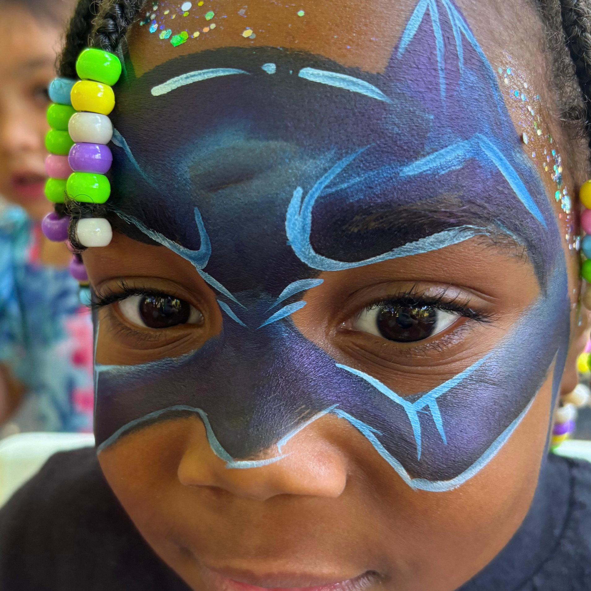 Close-up of a child's face with batman face paint, wearing multicolored bead earrings, with the focus on eyes and forehead.