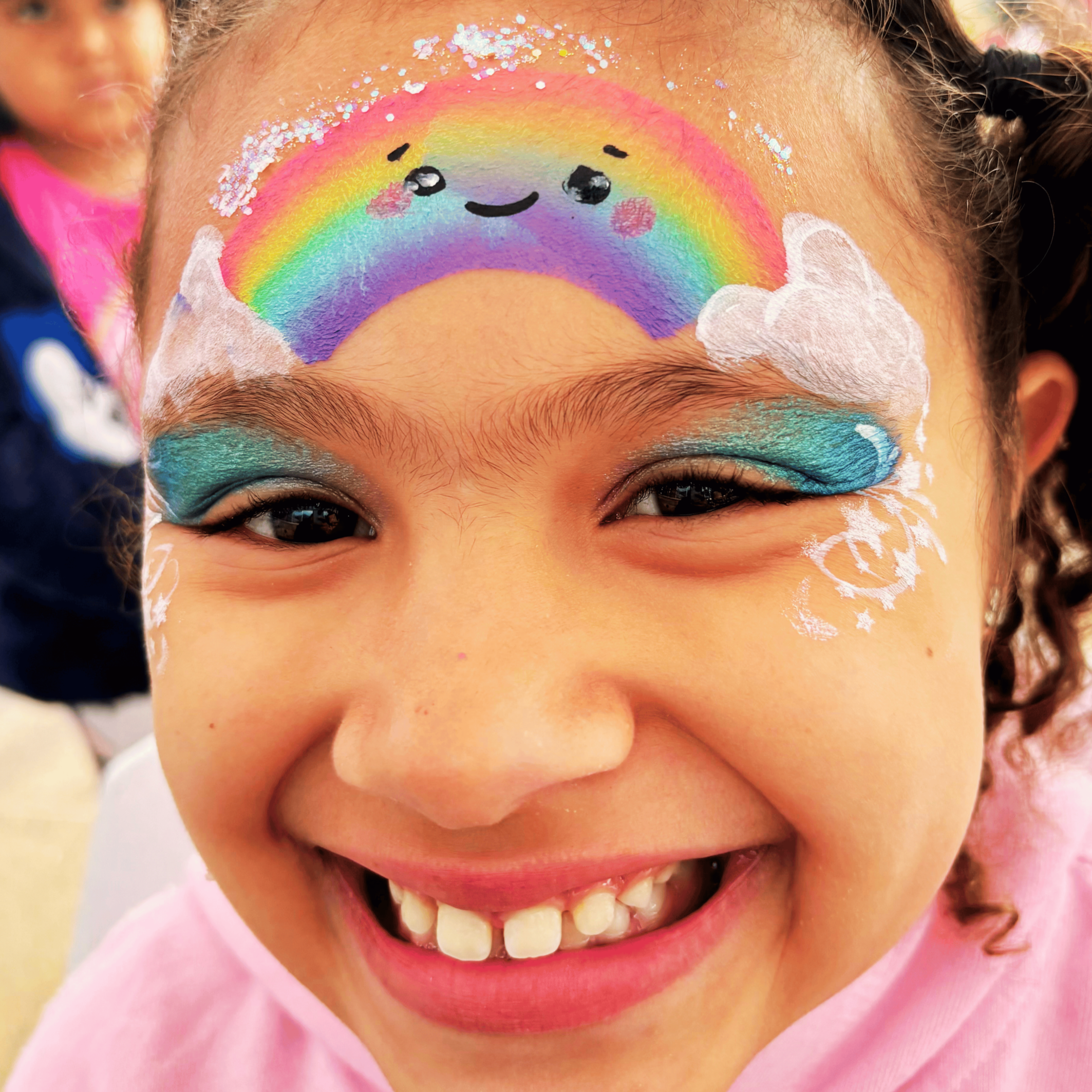 Close-up of a person's face with colorful rainbow and cloud face paint, smiling with visible teeth, and small glitter on the forehead and cheeks.