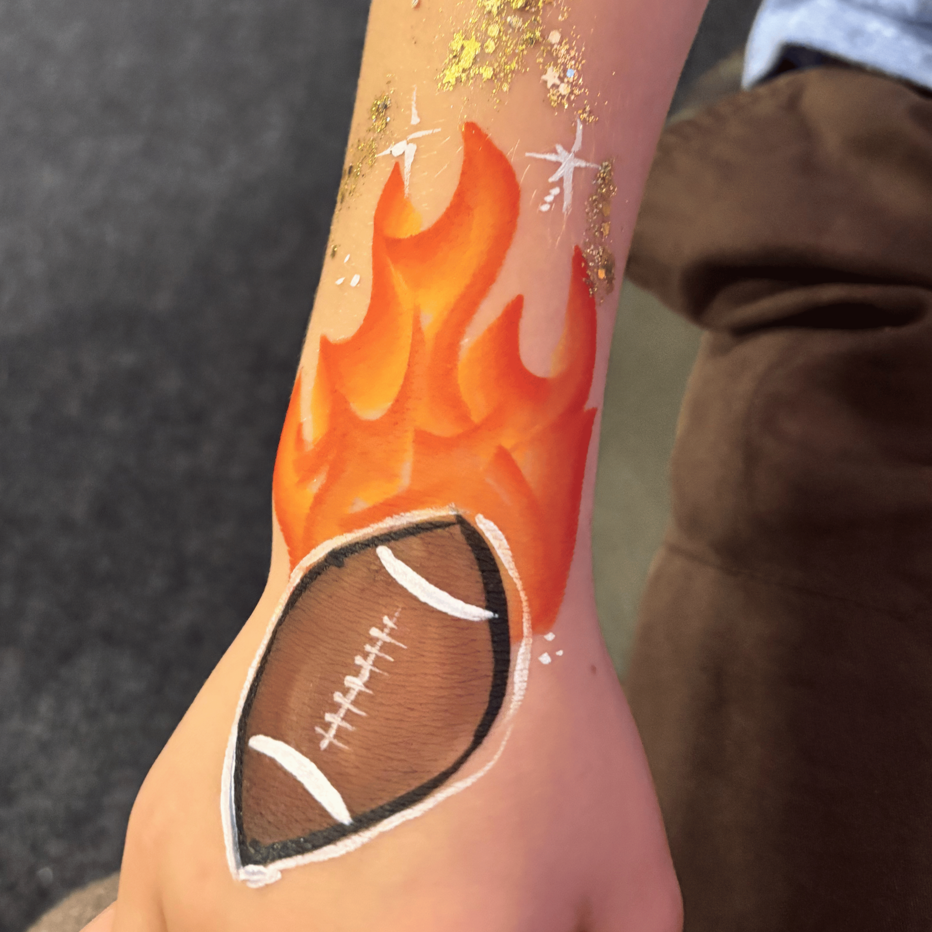 Close-up of a person's arm with a tattoo of a football designed with white laces and black outlines, decorated with golden glitter and white snowflake patterns.
