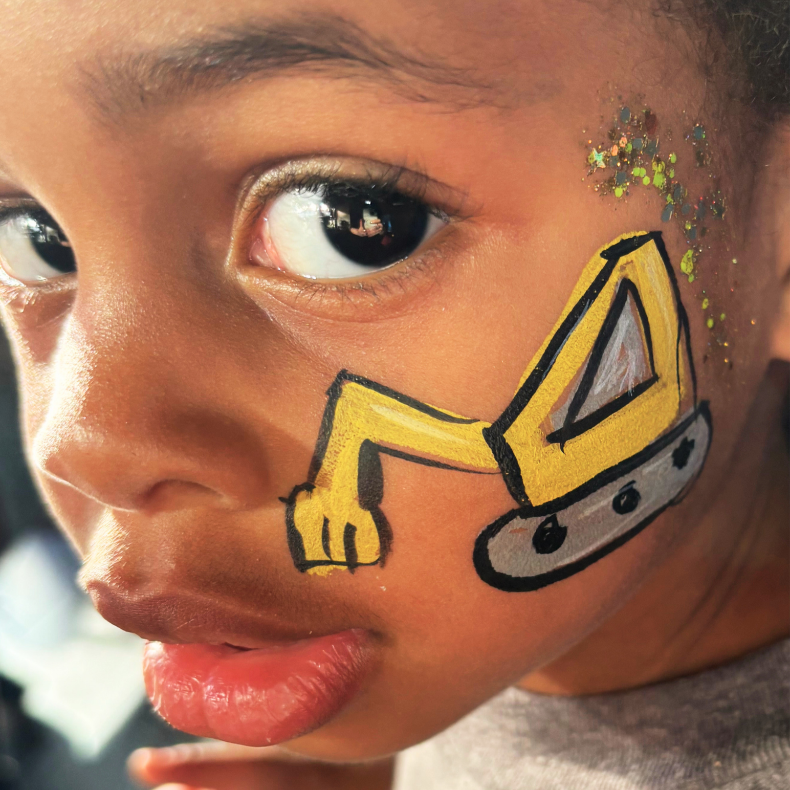 Close-up of a child's face with face paint featuring a yellow cartoonish construction vehicle and glitter near the cheek and eye.