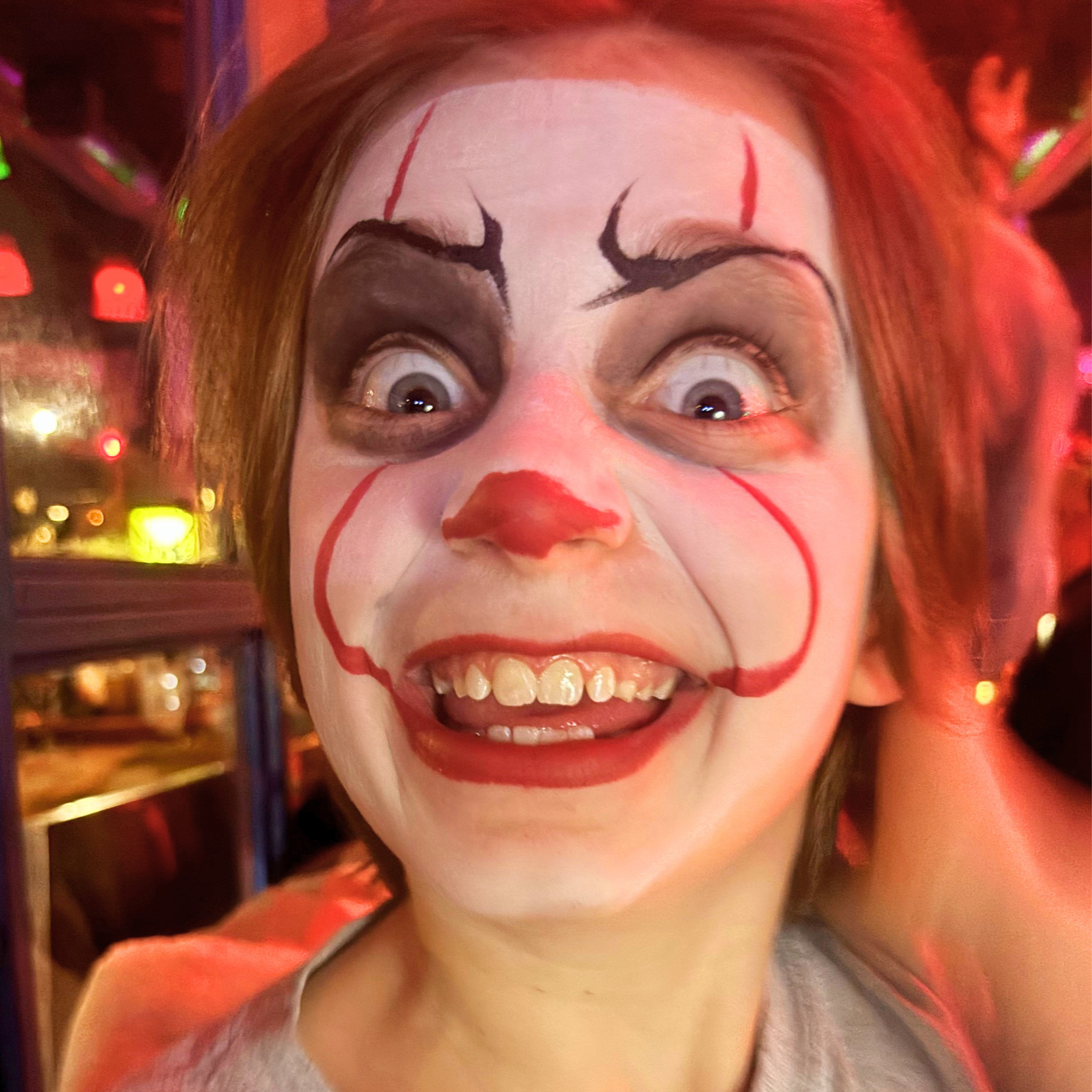 A person with face paint resembling a clown or mime, smiling with wide eyes, painted lips, and exaggerated makeup, in a setting with colorful blurry lights.