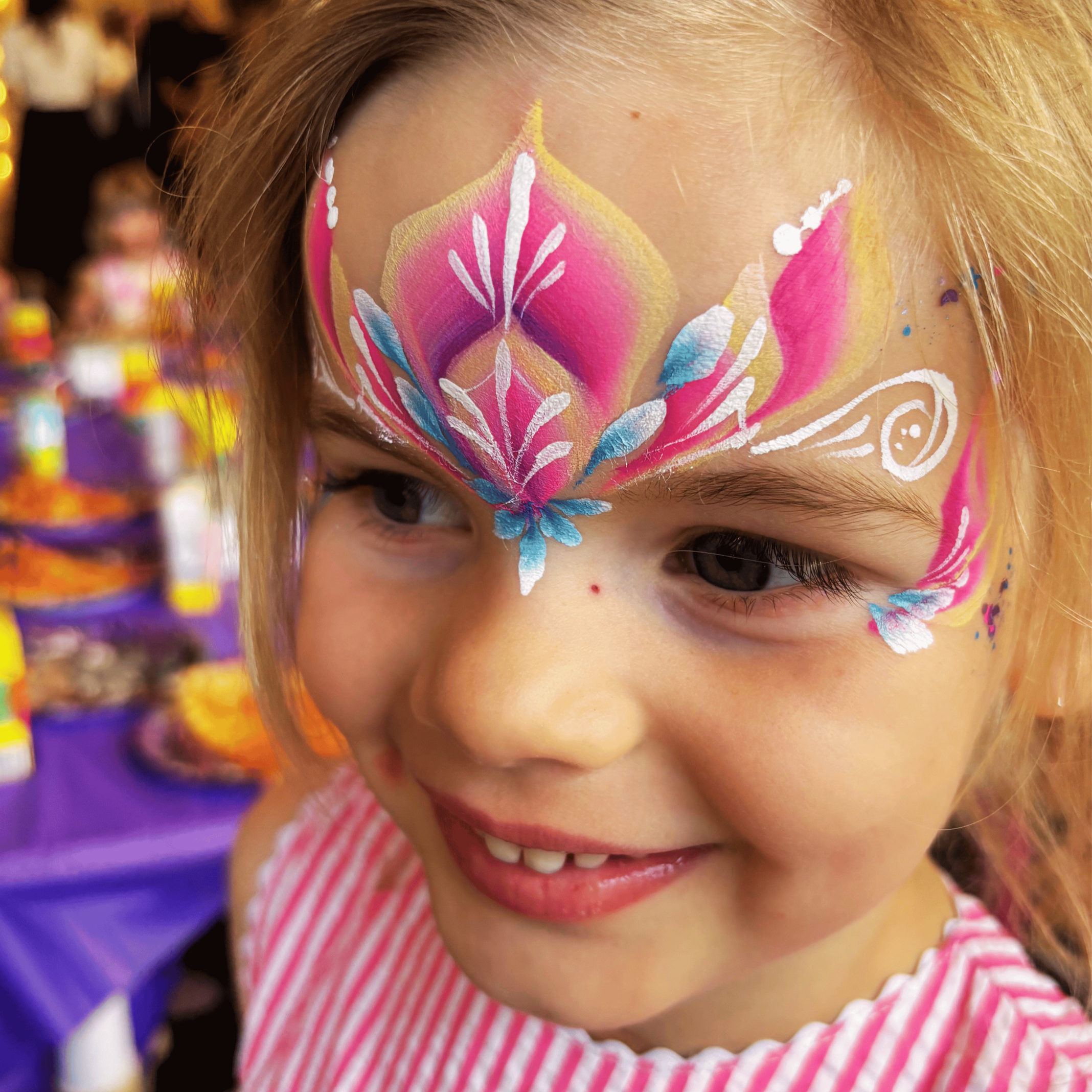 A person with face paint resembling a colorful butterfly with pink, yellow, blue, and white details, smiling and wearing a pink and white striped shirt.