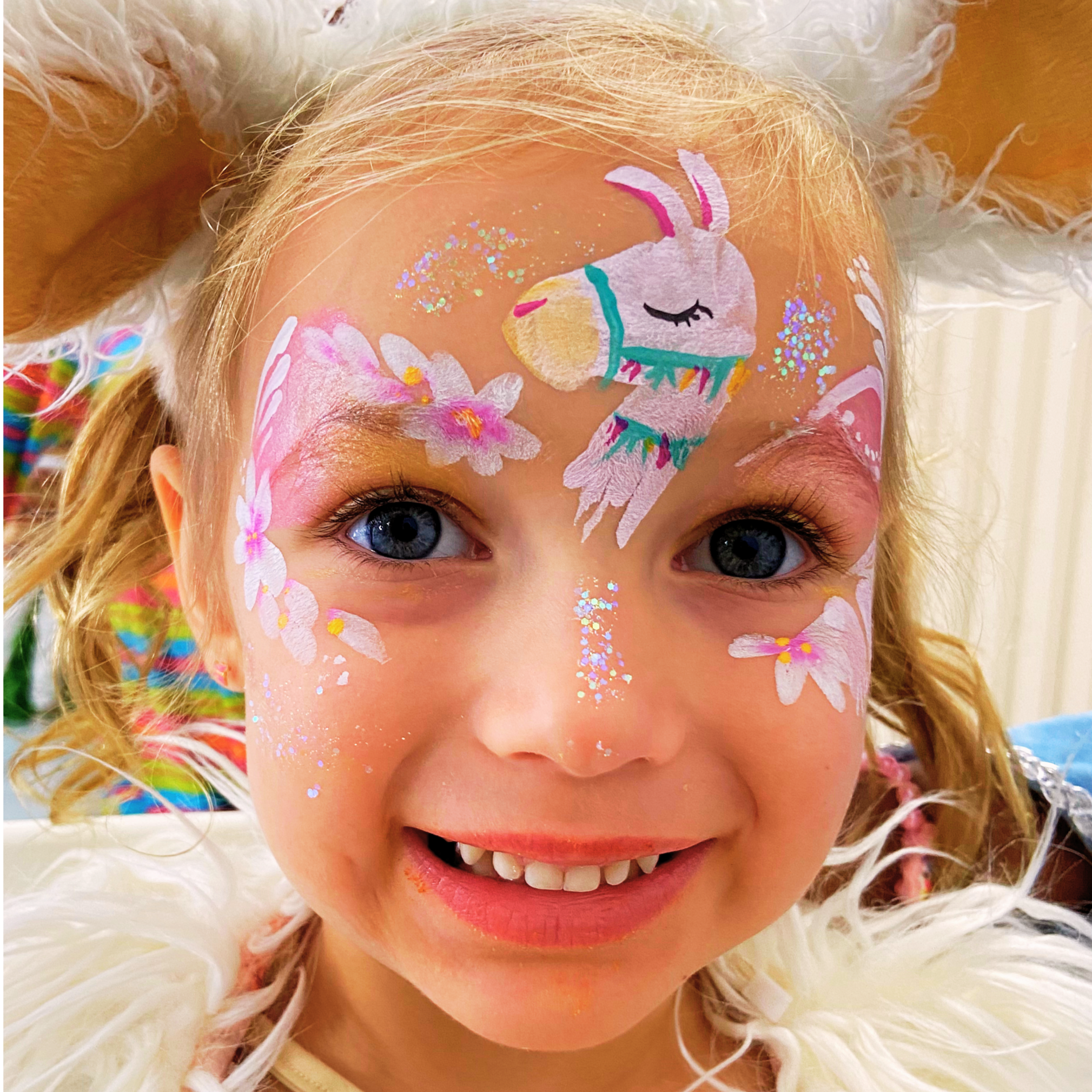 Close-up of a child's face with colorful face paint of a unicorn and flowers, with glitter and sparkles, smiling with teeth showing.
