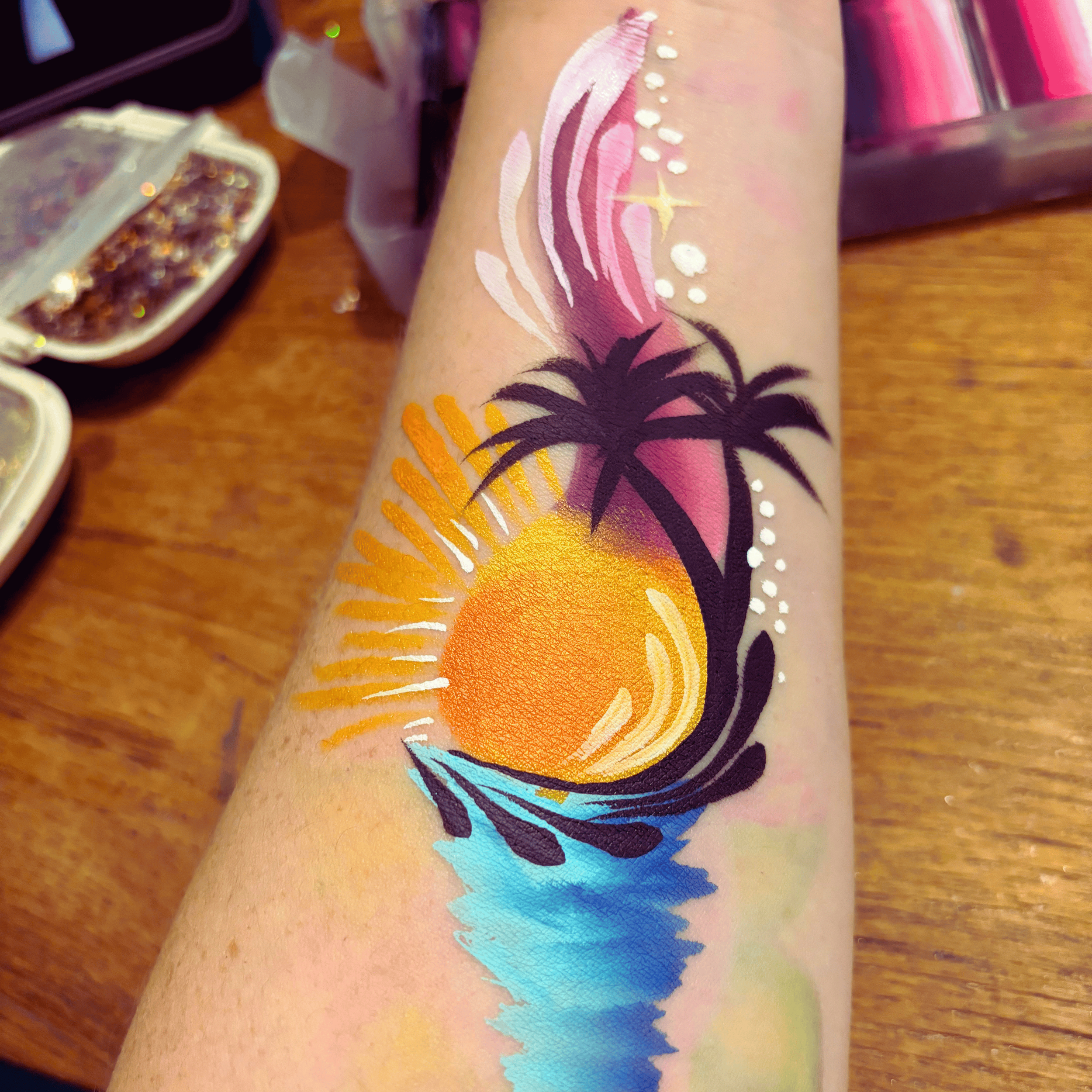 Close-up of a person's arm with detailed, colorful body paint featuring pink, purple, orange, yellow, and blue rainbow and palm tree designs.