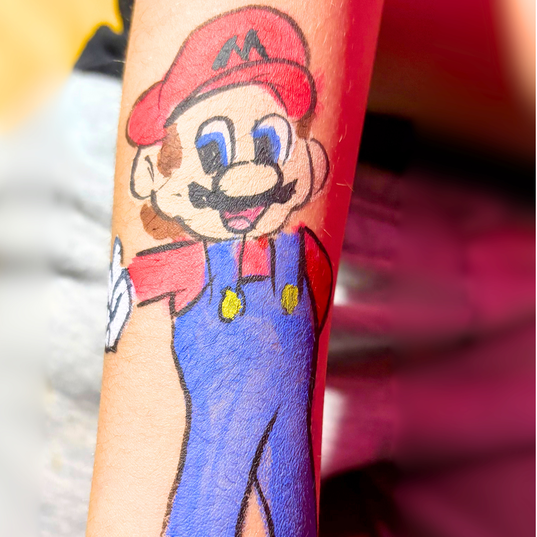 Colorful tattoo of Mario, a video game character, on a person's arm. Mario has a red hat with an 'M', blue overalls, and is smiling.