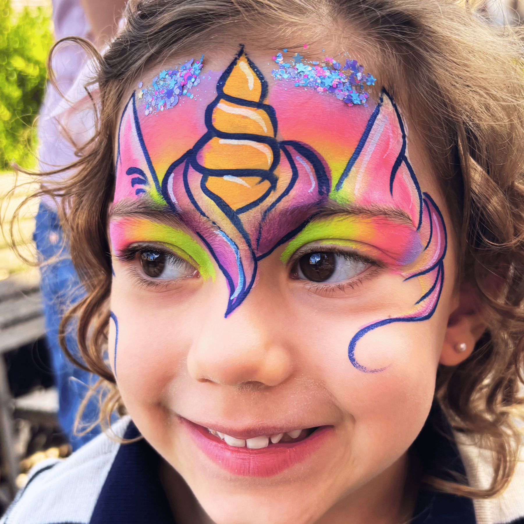 A child's face with colorful face paint, featuring a unicorn design with glitter around the forehead, pink, yellow, orange, and purple colors, and bold black outlines.