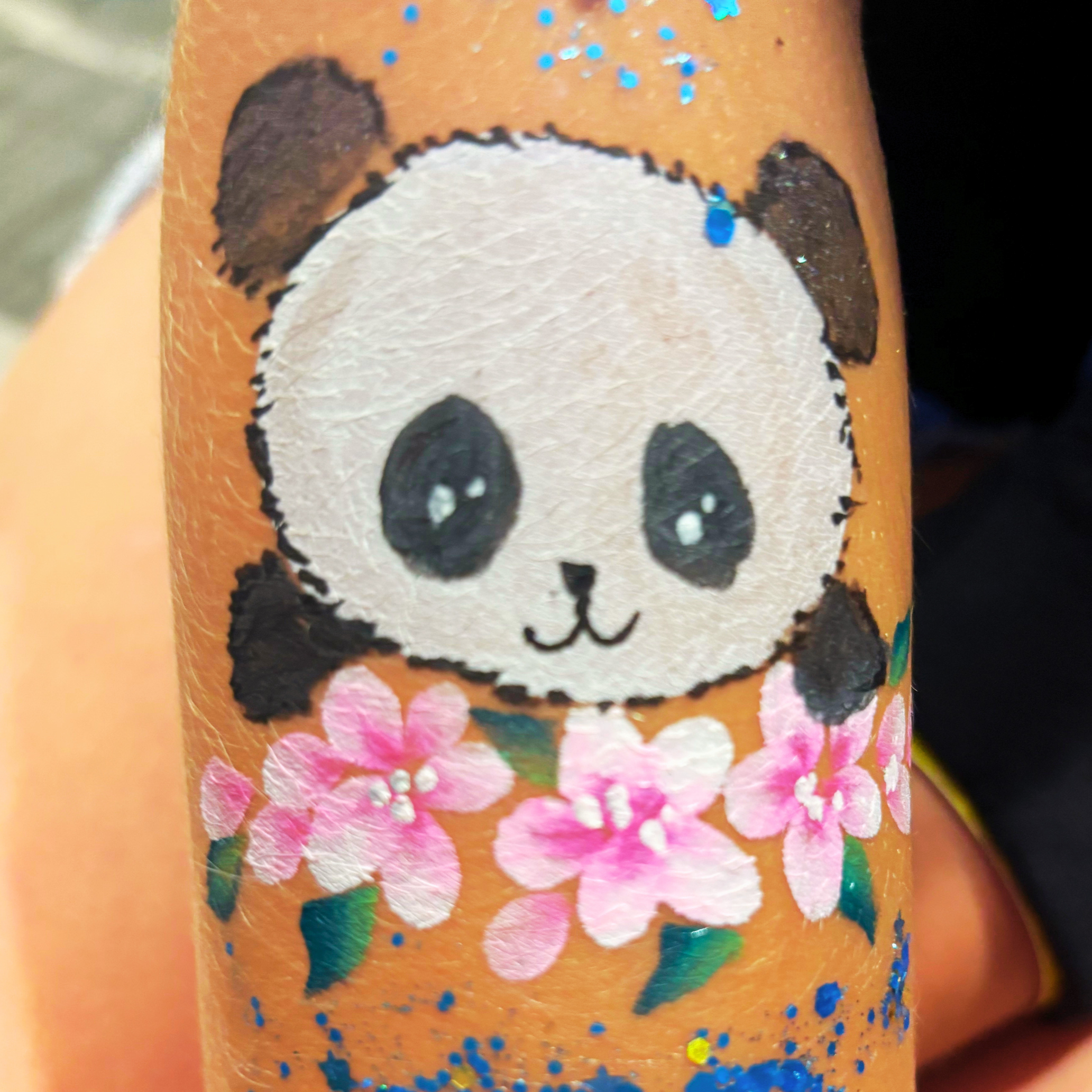 Close-up of a painted surface featuring a cartoon panda face with black ears, eyes, and nose on a white background, with colorful floral patterns and blue glitter dots surrounding it.