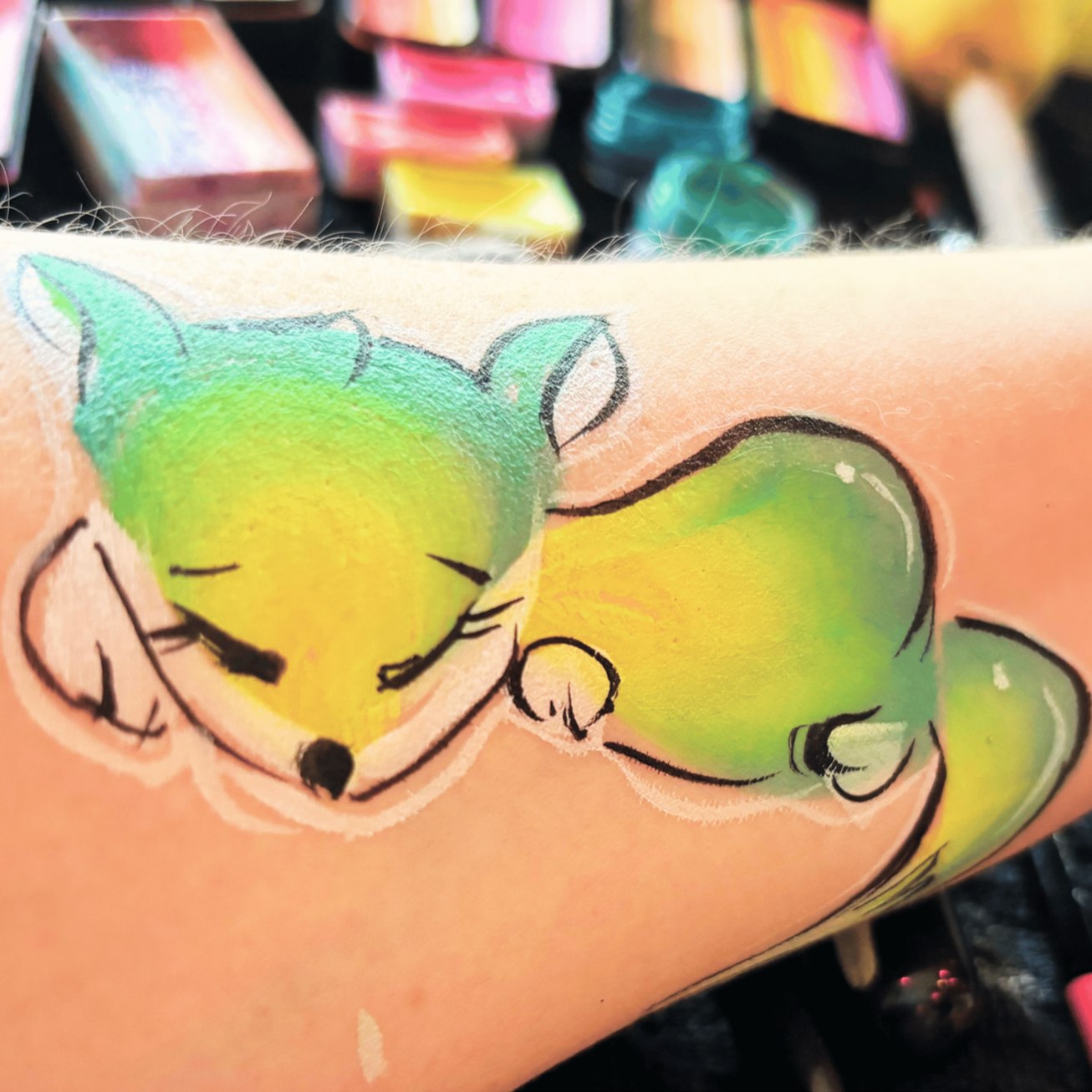 Tattoo of a sleeping fox with green and yellow colors on a person's arm, with art supplies blurred in the background.