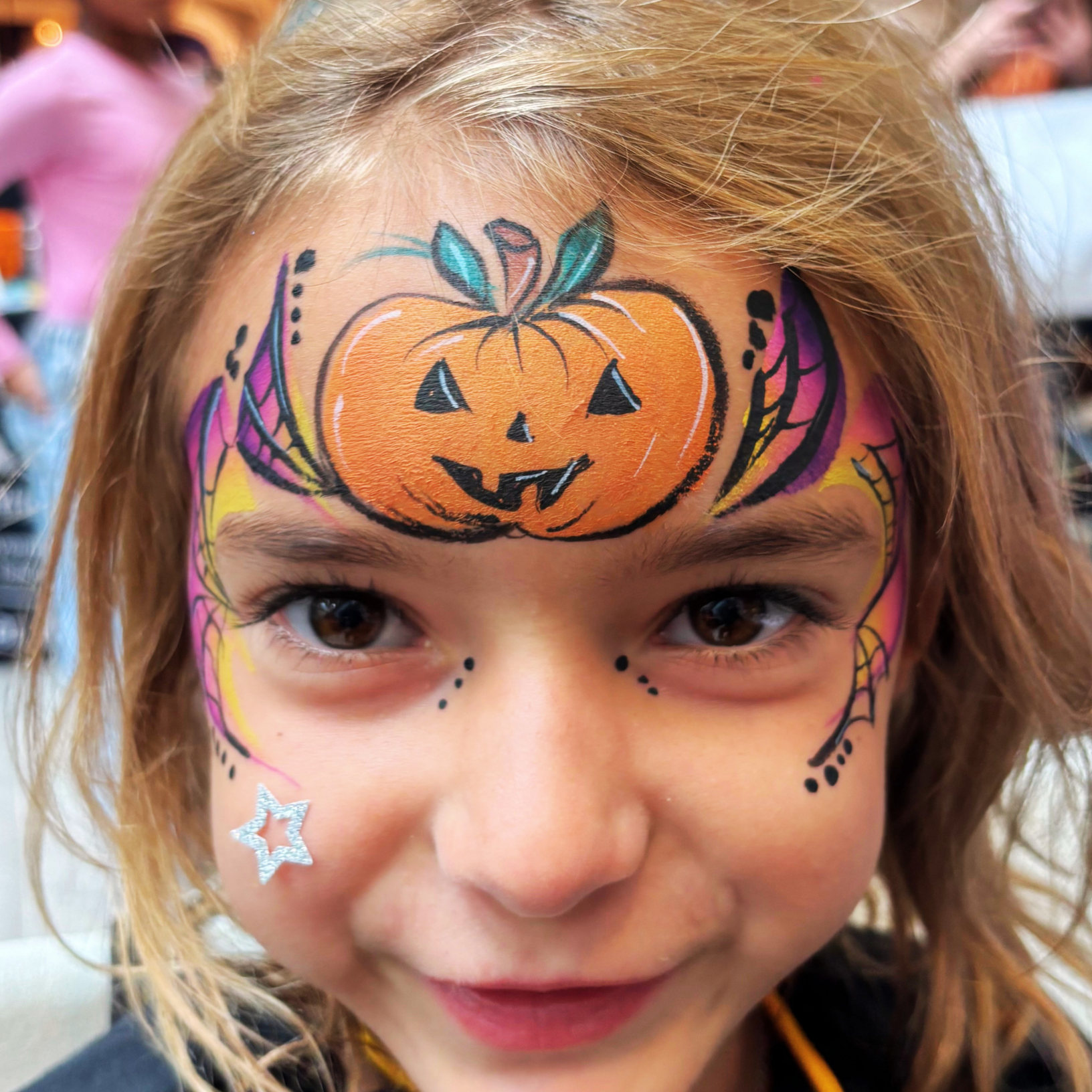 Close-up of a child's face with colorful halloween face paint featuring a pumpkin on the forehead, decorated with leaves and accents, and a star sticker on the cheek.