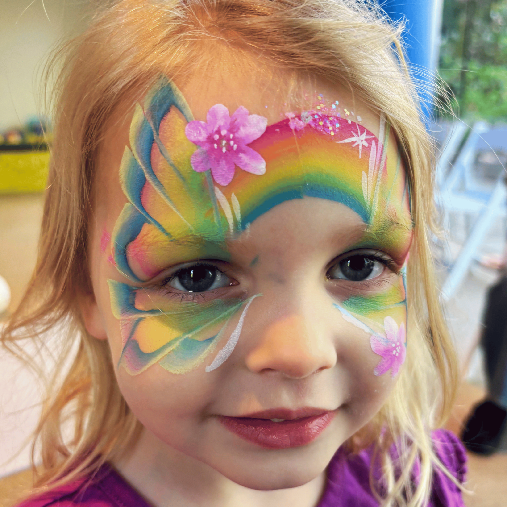 Close-up of a child's face with colorful rainbow and floral face paint covering the forehead, cheeks, and around the eyes.