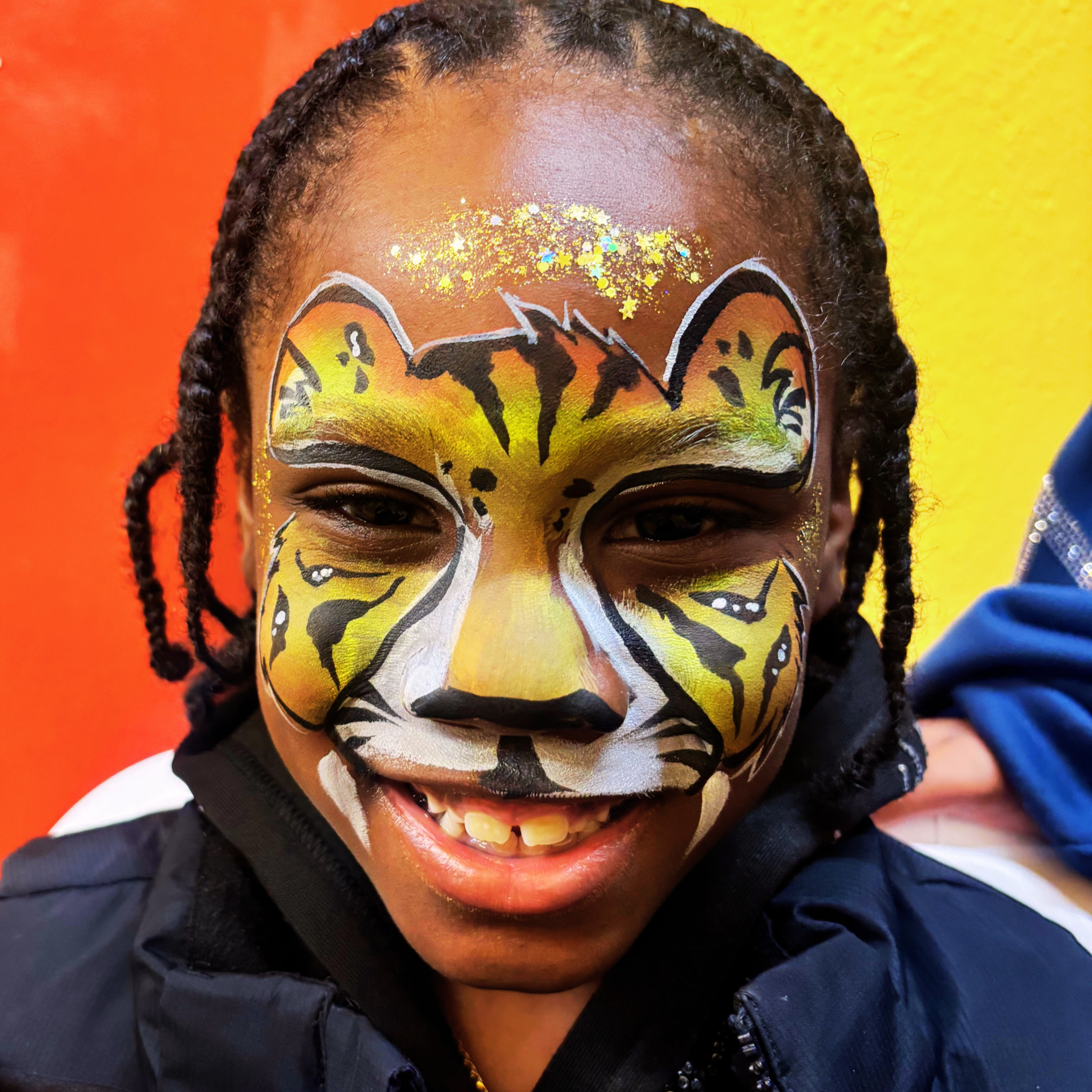Close-up of a person with a butterfly face paint design and glitter on the forehead, wearing a dark jacket, with an orange and yellow background.