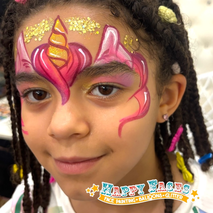 A young girl with face paint of a colorful unicorn, featuring pink, purple, and gold glitter details, on her forehead and face.