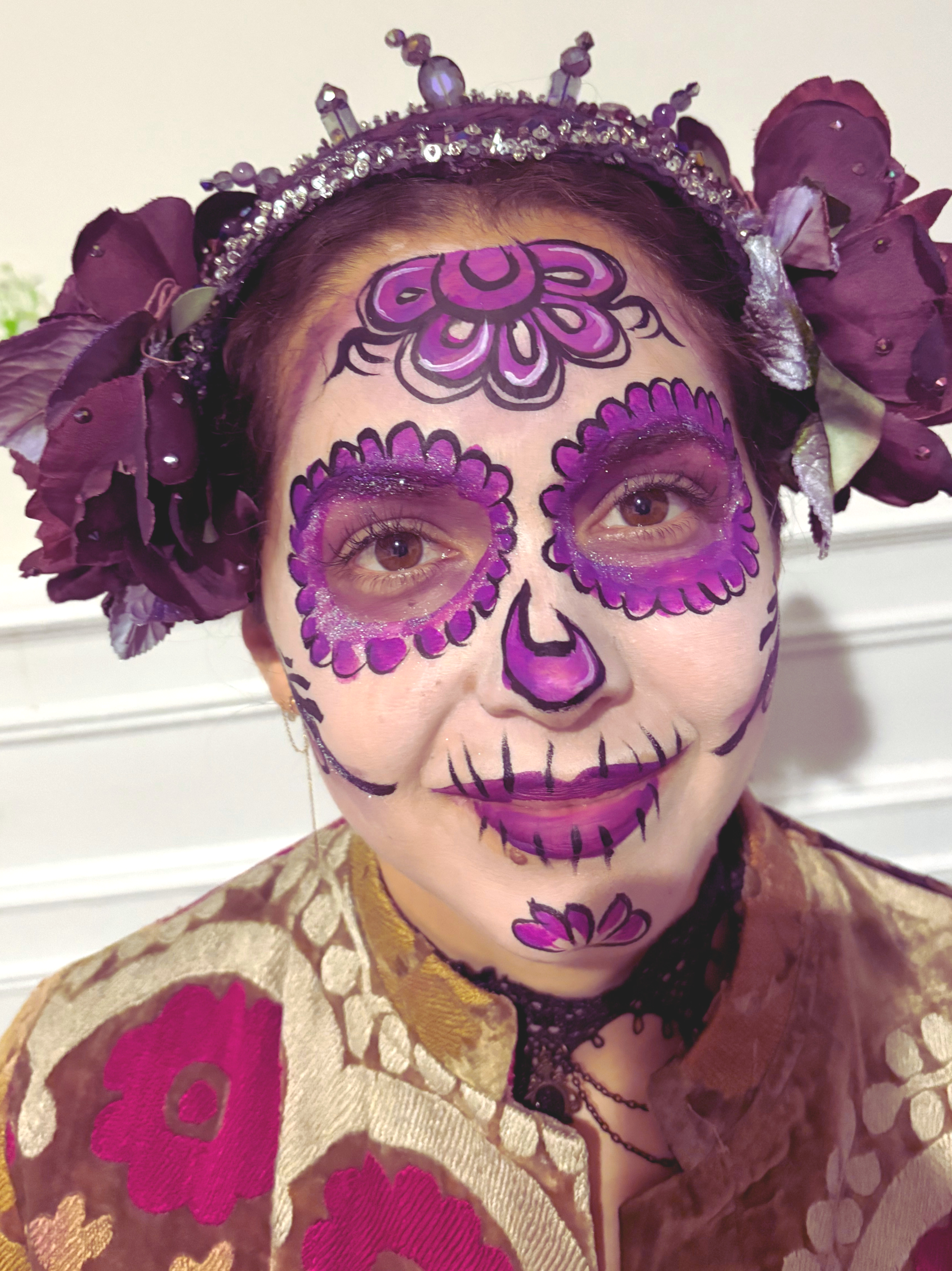 Person with face painted in the style of a sugar skull, featuring purple and black designs, surrounded by purple flowers and wearing a decorative headpiece with beads and feathers.