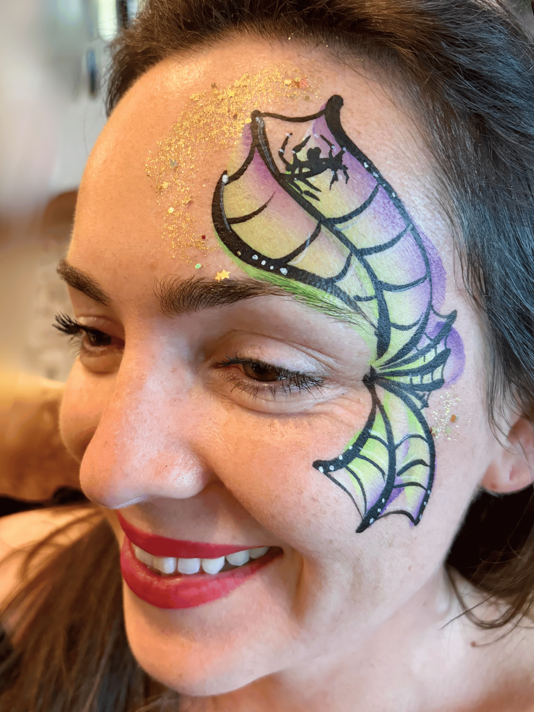 A woman with face paint in the shape of a butterfly with purple, yellow, and green colors, gold glitter on her forehead, and red lipstick smiling.
