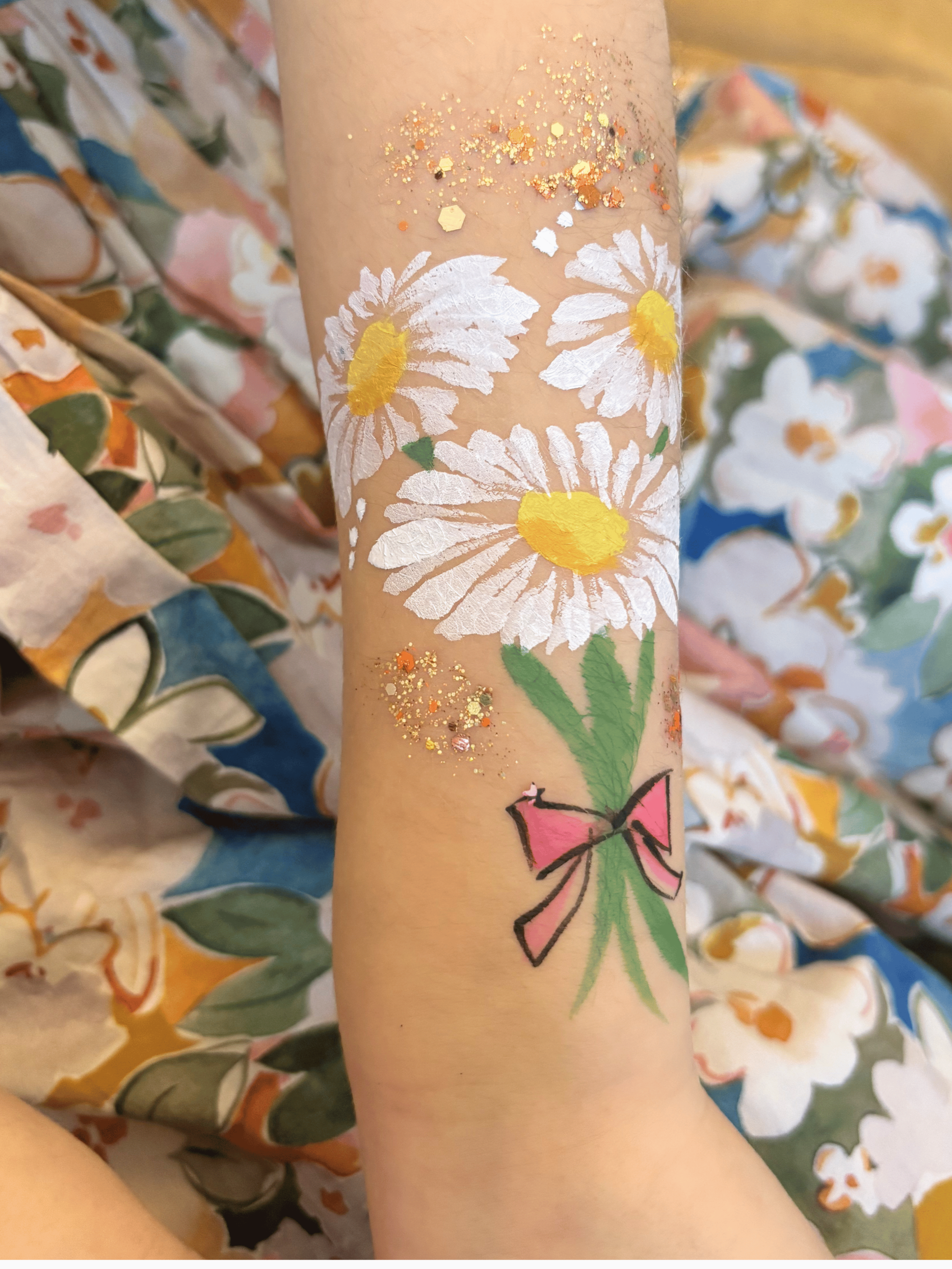 Temporary tattoo of three white daisies with yellow centers and green stems, with gold glitter underneath, on a person's forearm. Floral patterned fabric in the background.