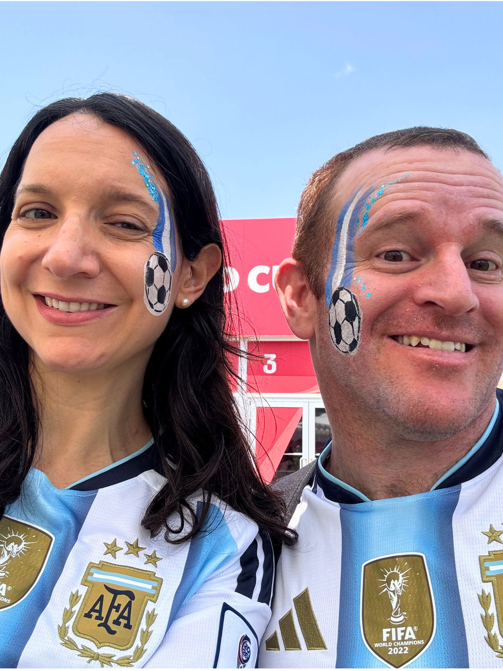 Two people wearing Argentina soccer jerseys with face paint, celebrating at the 2022 FIFA World Cup, with a blue sky in the background.