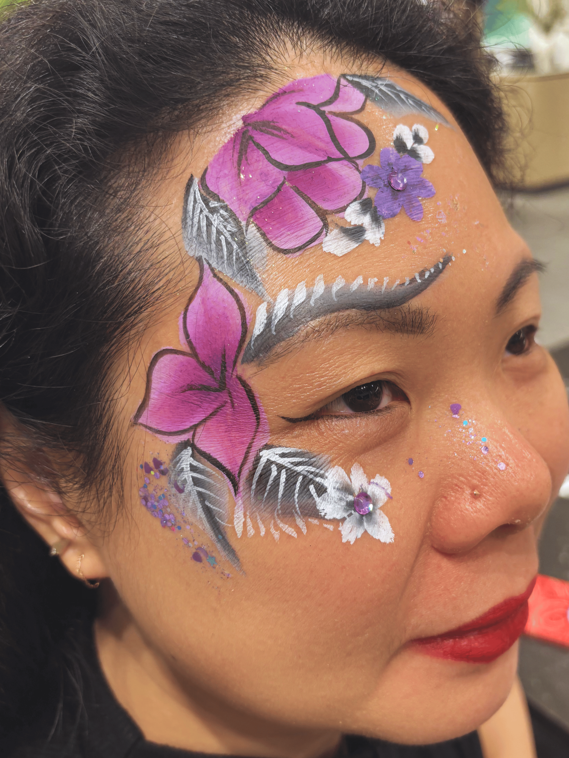 Close-up of a woman's face with colorful butterfly and floral face paint, including pink and purple butterflies, white and purple flowers, black and white leaves, and glitter accents.