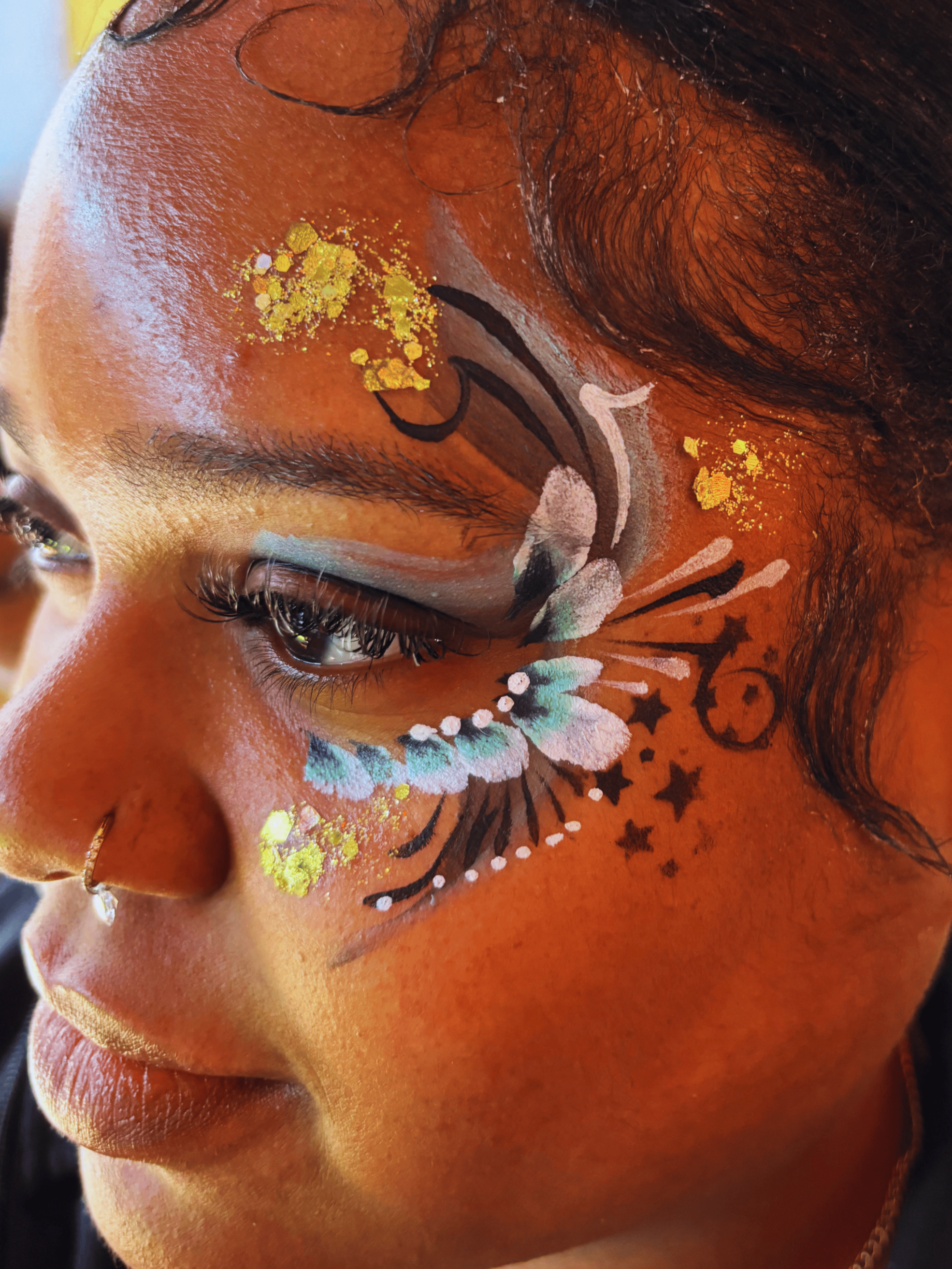 Close-up of a person's face with intricate face paint and glitter, including paisley embellishments on the nose and elaborate designs around the eye.