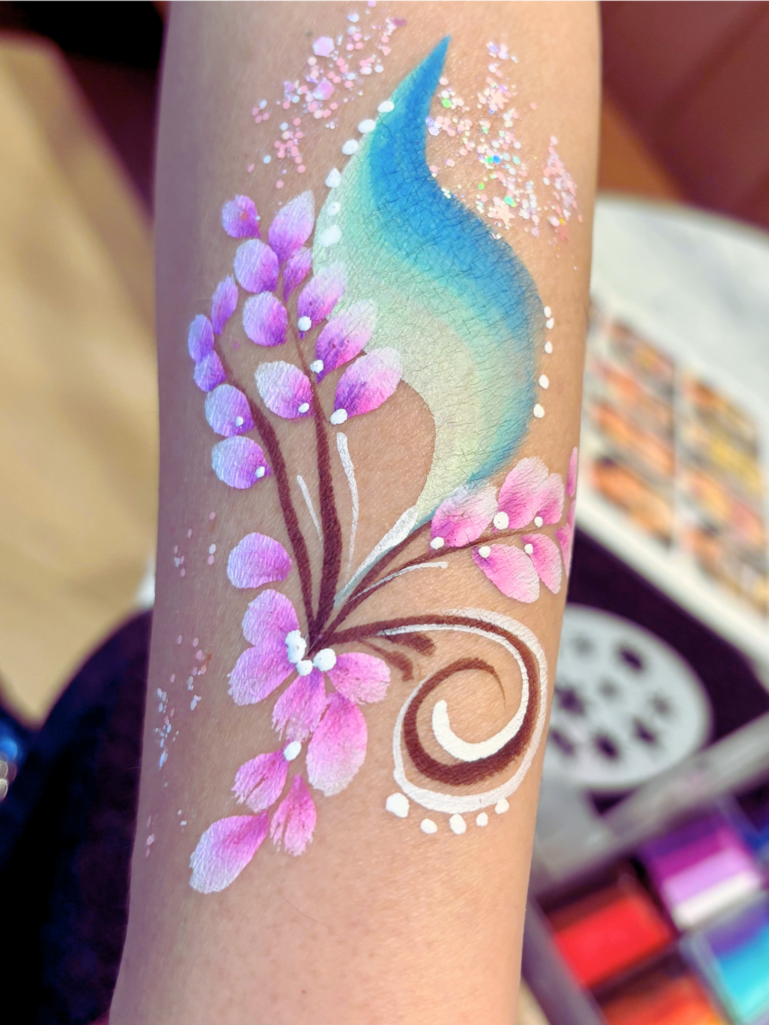 Close-up of a colorful body art or temporary tattoo featuring pink and purple flowers with white accents, a blue and green leaf, white decorative elements, and glitter, on a person's arm.
