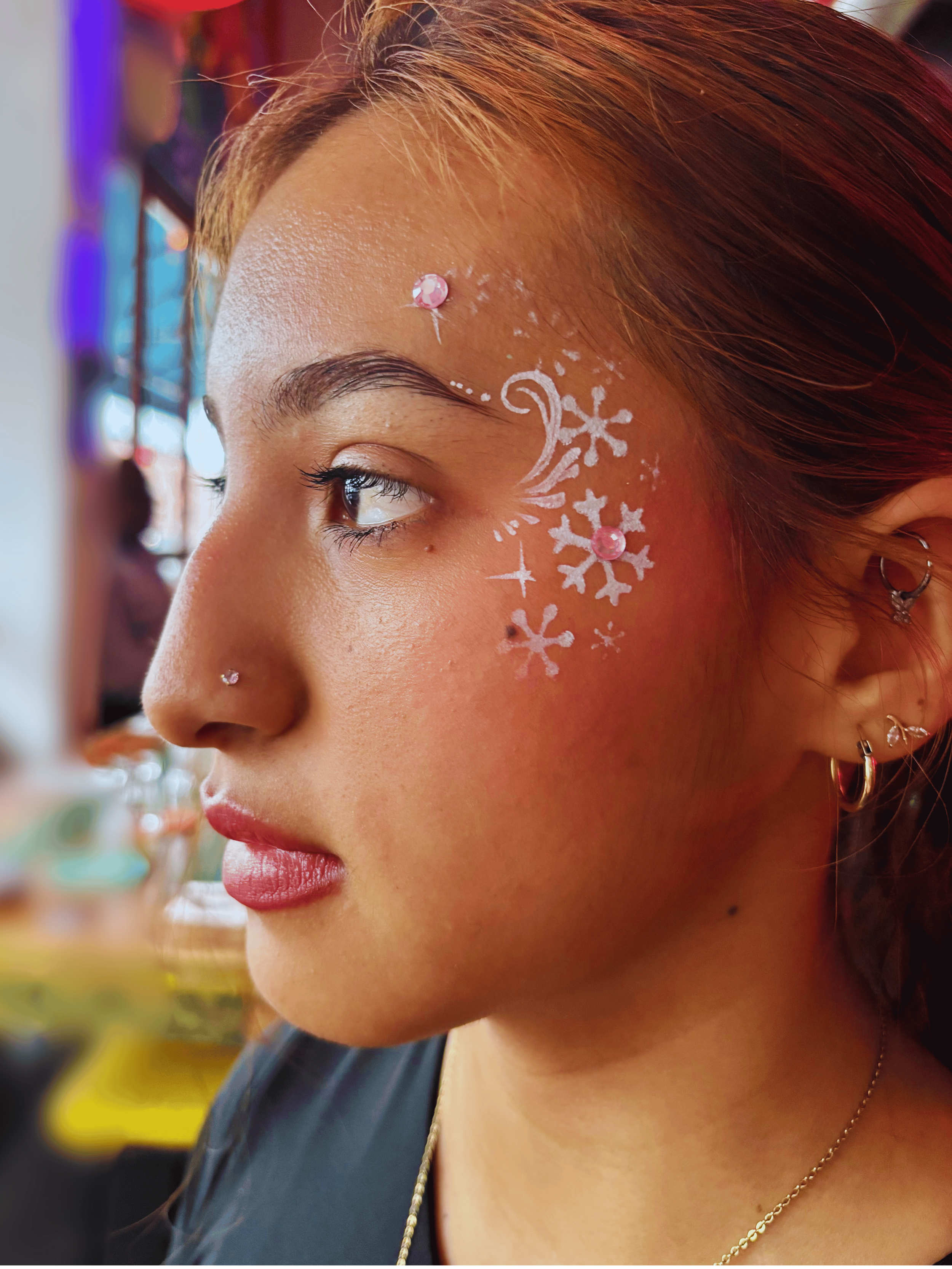 Close-up of a woman with red hair and makeup decorated with white snowflake patterns and pink rhinestones on her face, wearing earrings and a gold necklace.