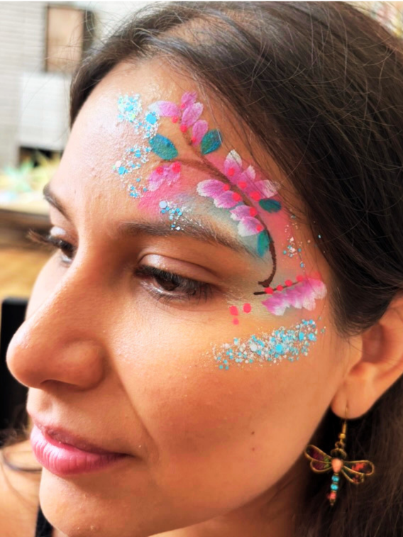 Close-up of a woman with detailed face paint featuring pink cherry blossoms and blue accents on her temple and cheek.