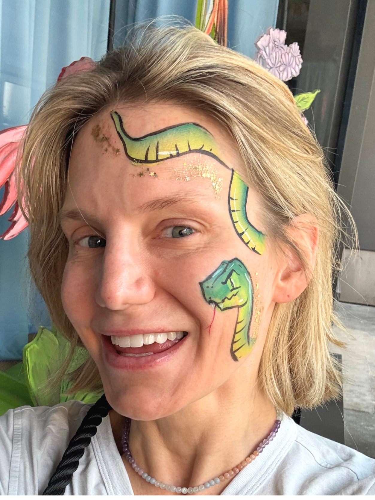 Woman with face painted as a green snake with yellow accents and black outlines, gold glitter around her eye, smiling with teeth showing, wearing white shirt and pearl necklace.