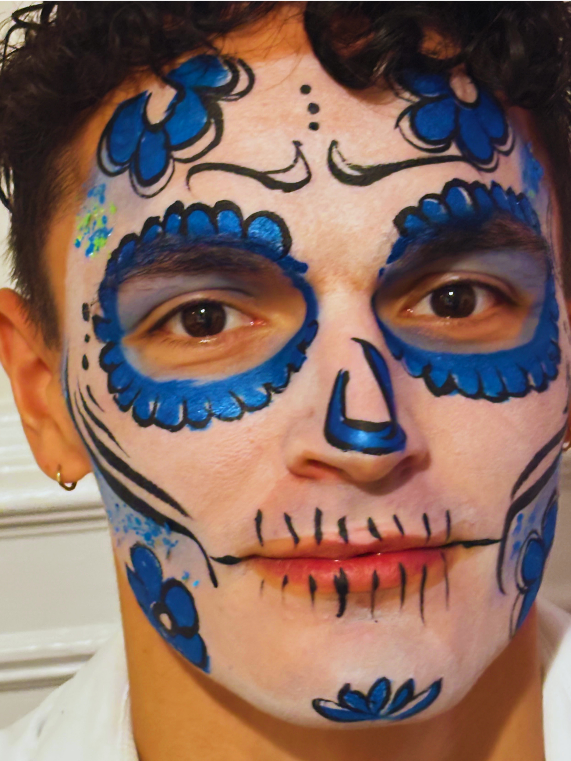 Person's face painted with a colorful sugar skull design featuring blue, black, and pink colors, with floral and decorative patterns.