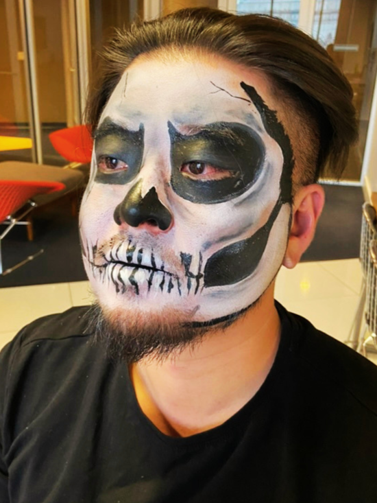 Man with skull face paint in an indoor setting, wearing a black shirt, with chairs and windows in the background.