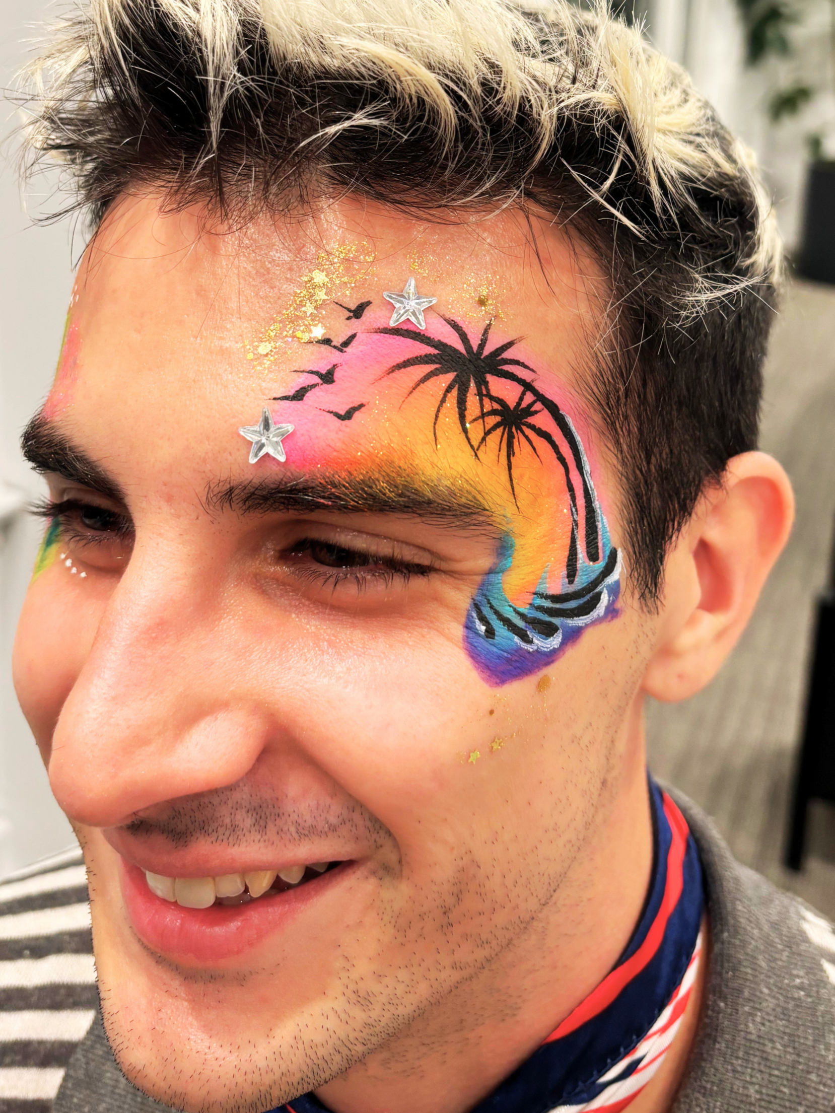 Close-up of a smiling young man with black and blonde hair, colorful face paint featuring a sunset, silhouette palm trees, birds, pink, orange, blue, and purple hues, gold glitter, and star-shaped embellishments.