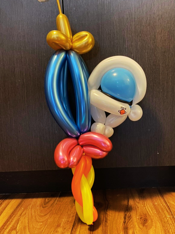 A colorful balloon sculpture of a flower with yellow, orange, pink, blue, white, and beige balloons, attached to a dark wall and wooden floor.