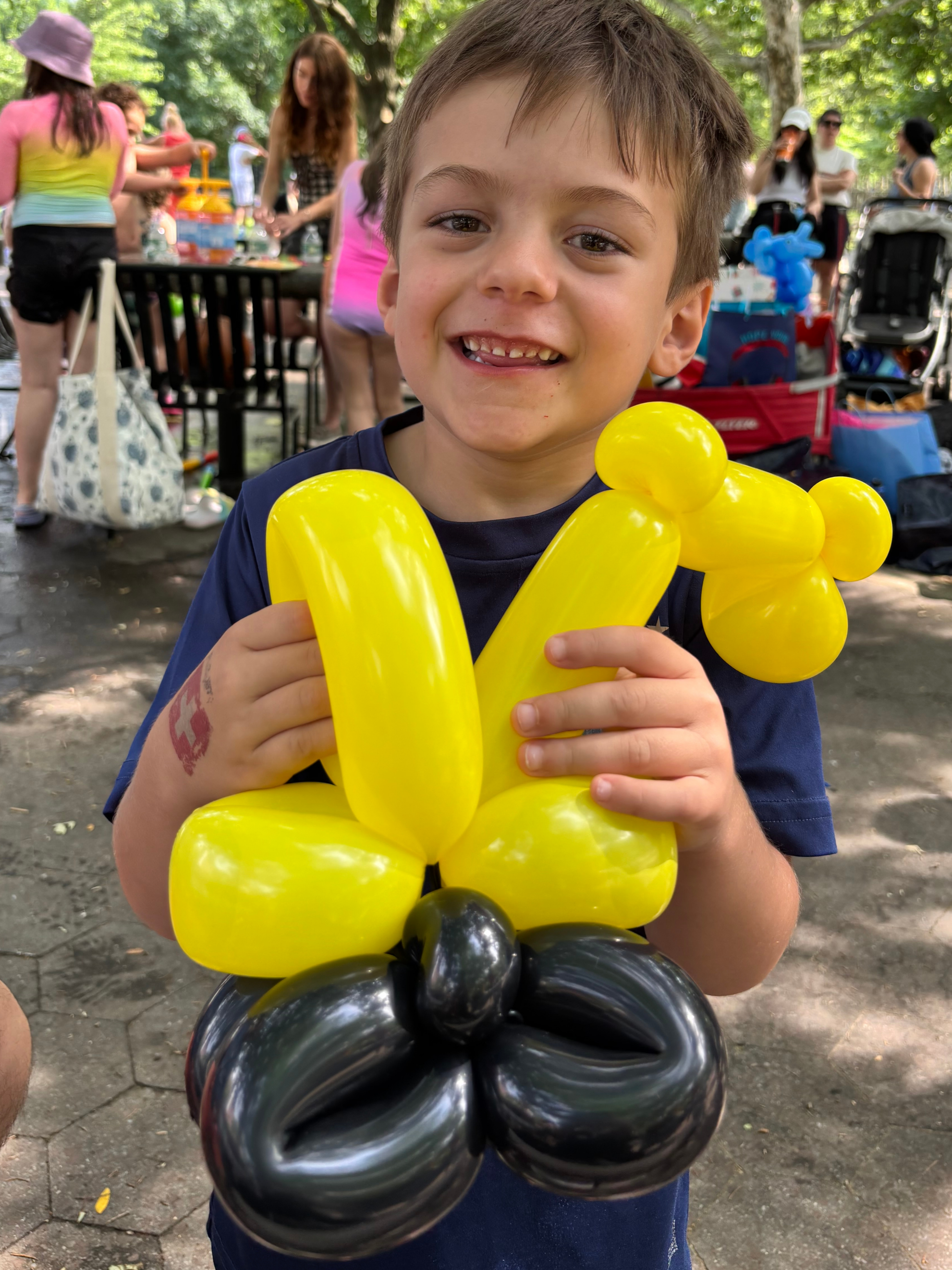 A young boy smiling while holding a yellow balloon animal at an outdoor event with trees and people in the background.