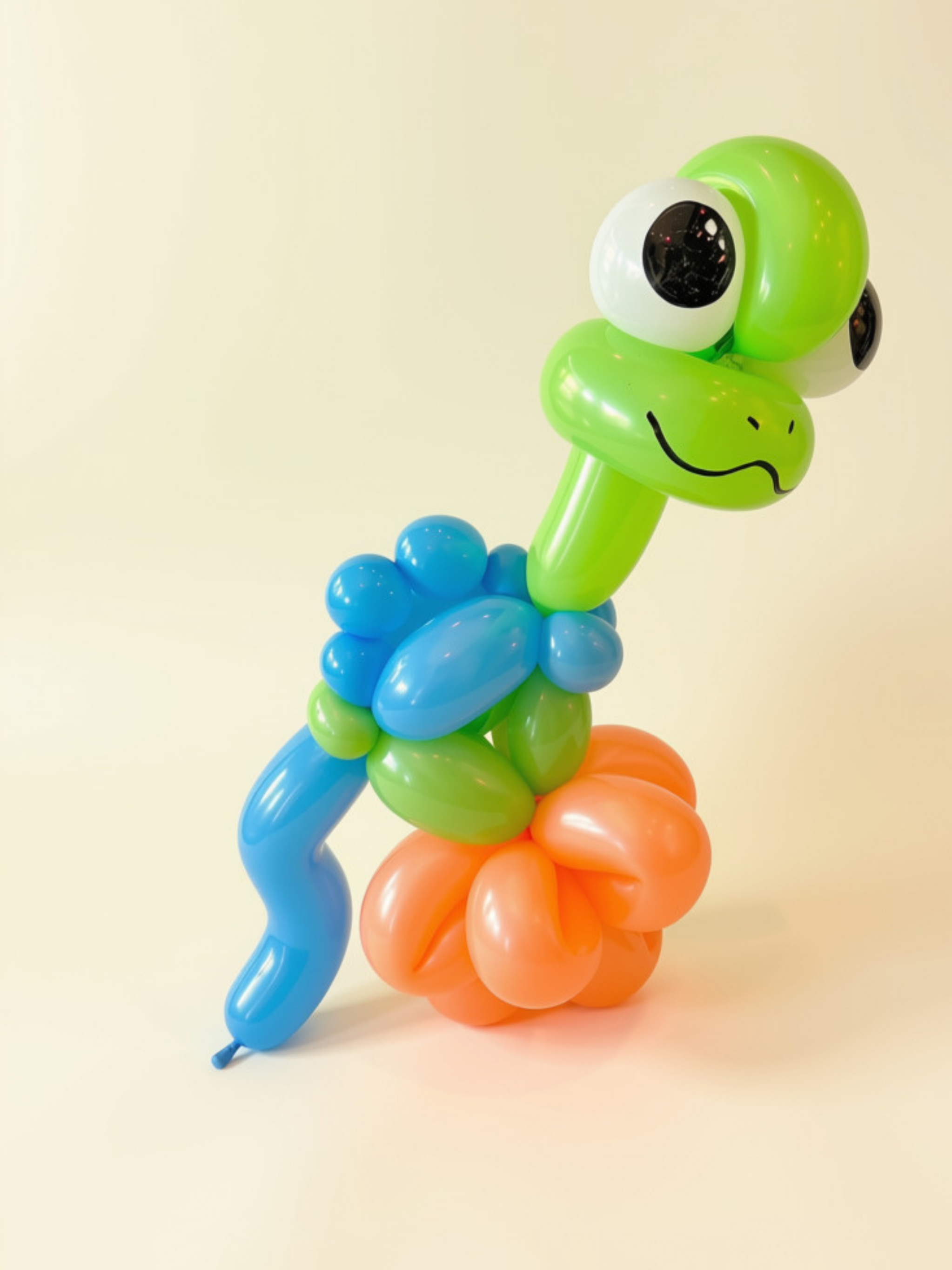 Colorful balloon sculpture resembling a whimsical animal with a green face, large eyes, and a smiling mouth, standing on a flower-shaped base made of orange balloons, against a plain off-white background.