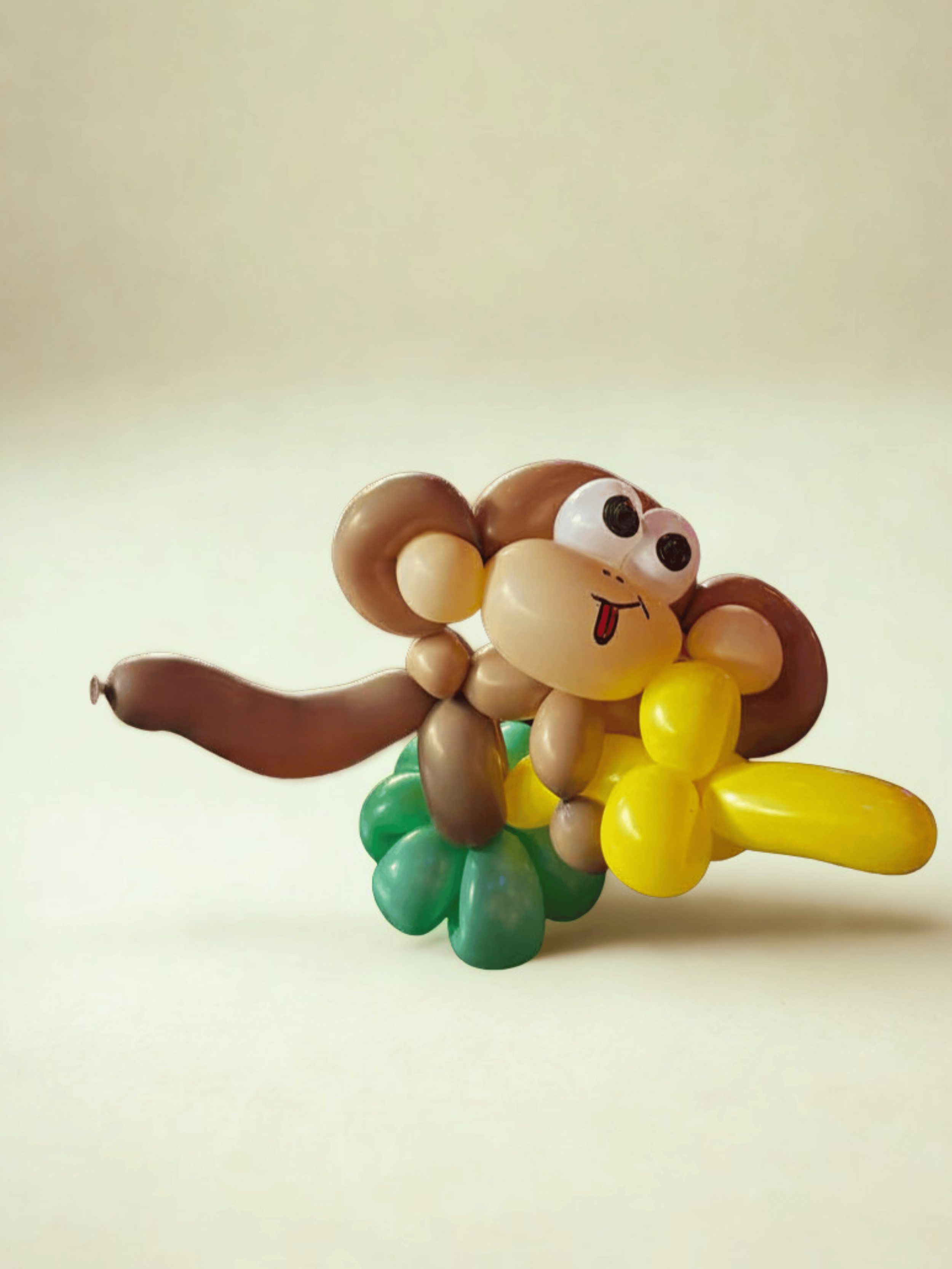 Balloon sculpture of a cartoon-style monkey with a smiling face, big eyes, sticking out tongue, sitting on a colorful cluster of balloons resembling flowers.