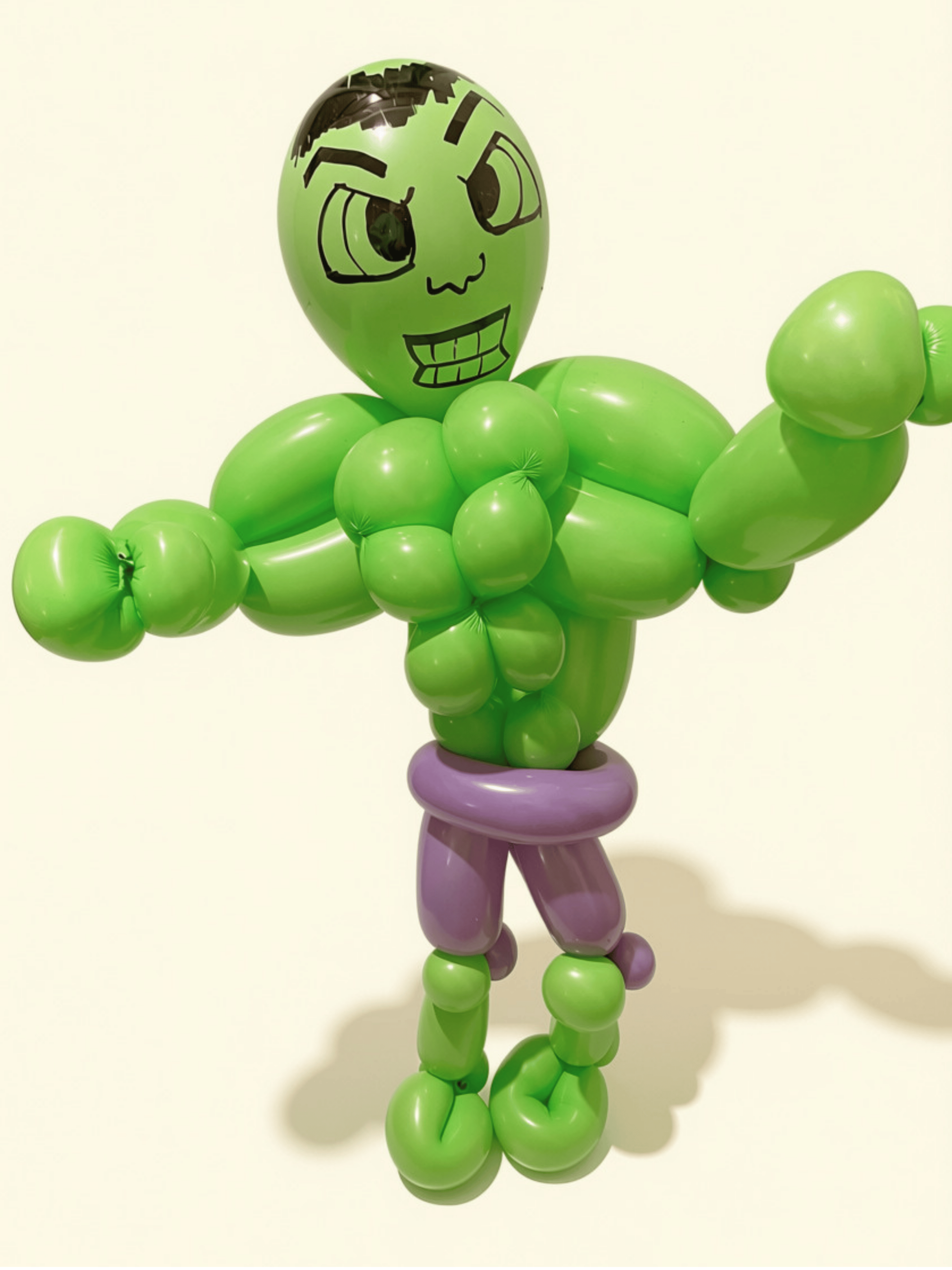Balloon sculpture of a cartoon-style humanoid figure with a green head, large eyes, and a wide smile, raised arm in a flexing pose, standing on a purple base against a plain white background.
