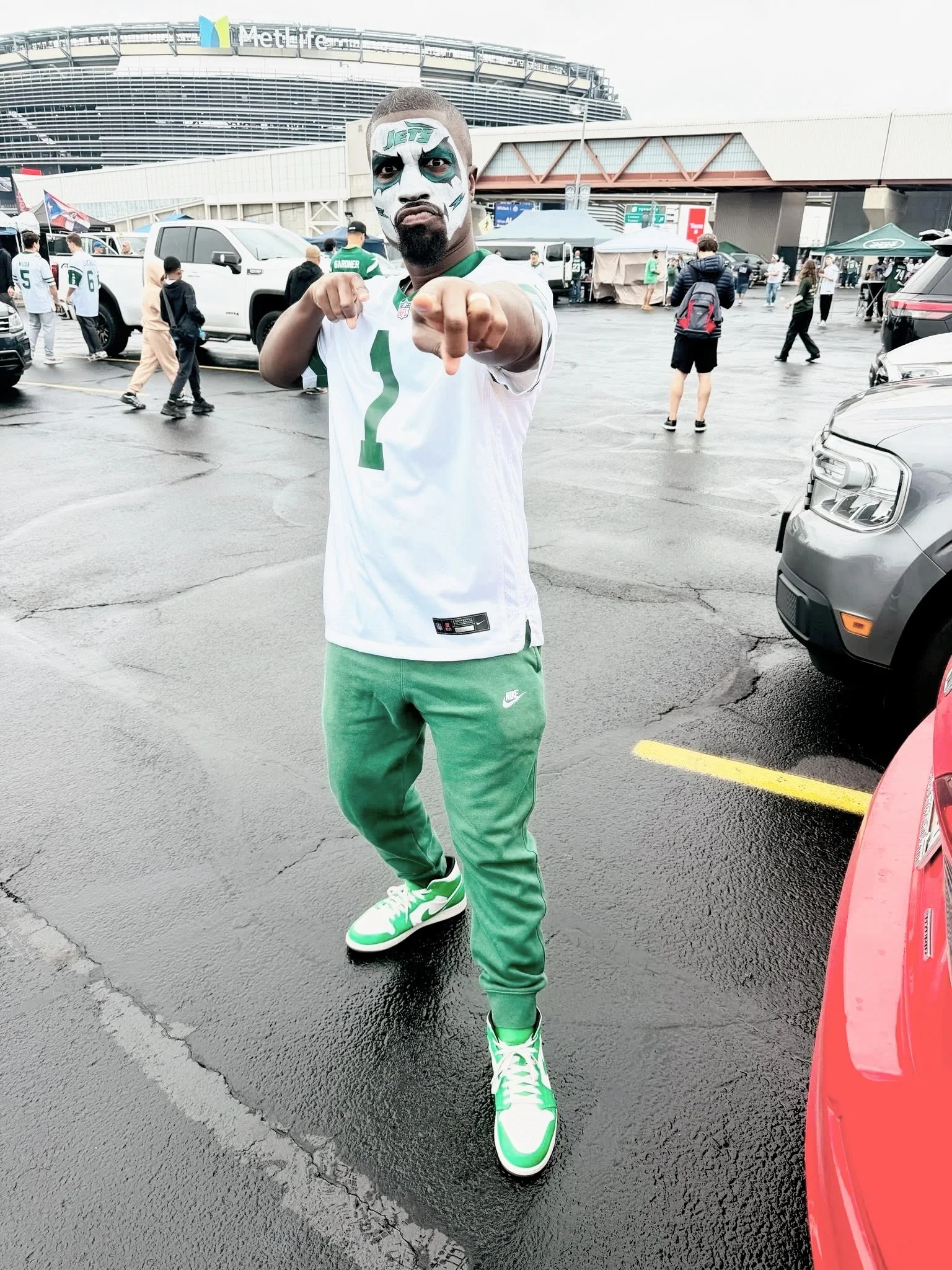 A man with face paint resembling the New York Jets logo, pointing at the camera, standing in a parking lot outside MetLife Stadium, wearing a white Jets jersey, green sweatpants, and green and white sneakers.