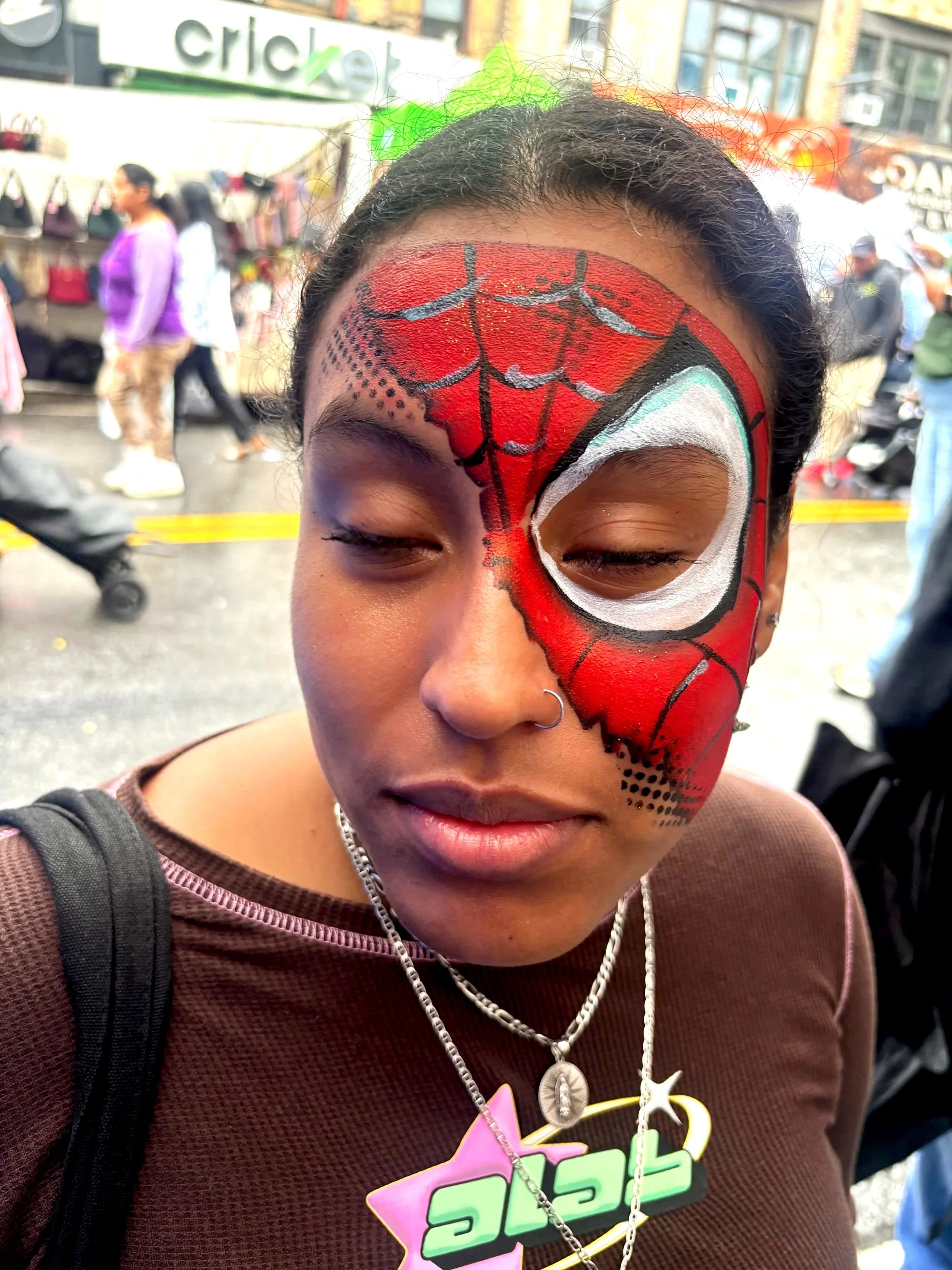 A person with half of their face painted as Spider-Man, with red, black, and white colors. They are wearing a necklace with a shell pendant and a neon-colored necklace, and have a nose ring. The background shows other people and a street scene.