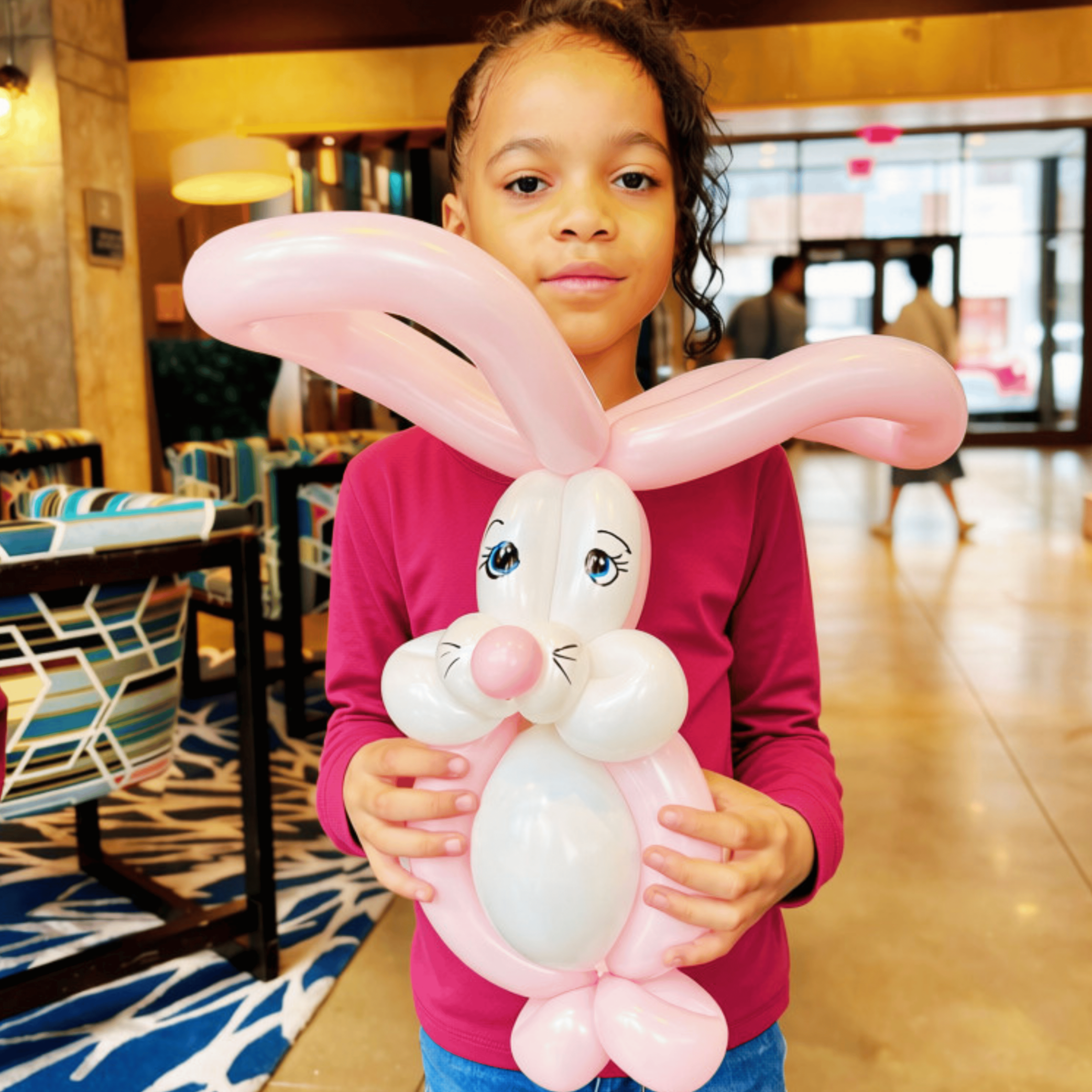Young girl holding pink balloon sculpture of a bunny with a smiling face inside a store or mall.