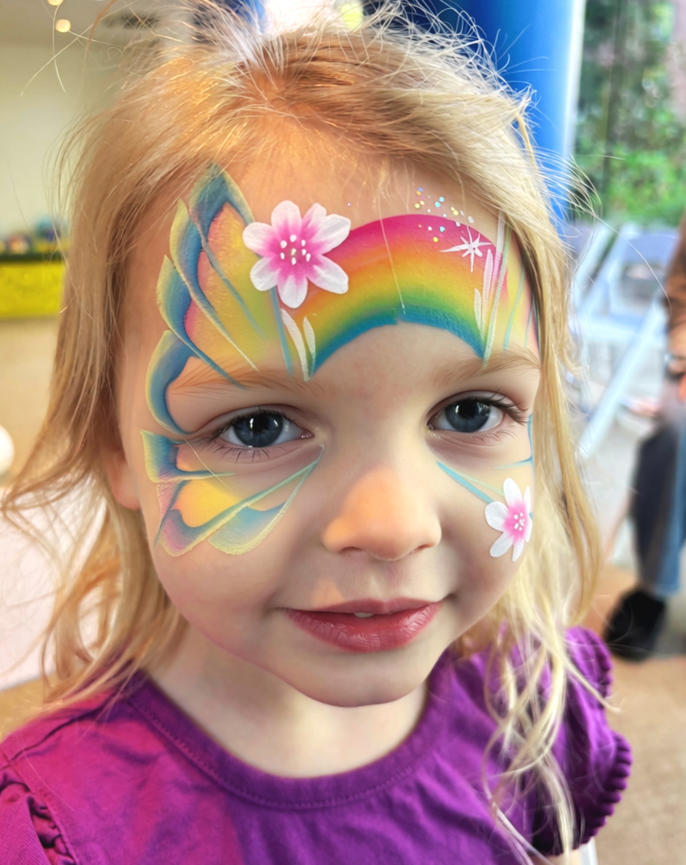 A young girl with blonde curly hair has colorful rainbow and flower face paint on her forehead and cheeks, and is wearing a purple shirt.