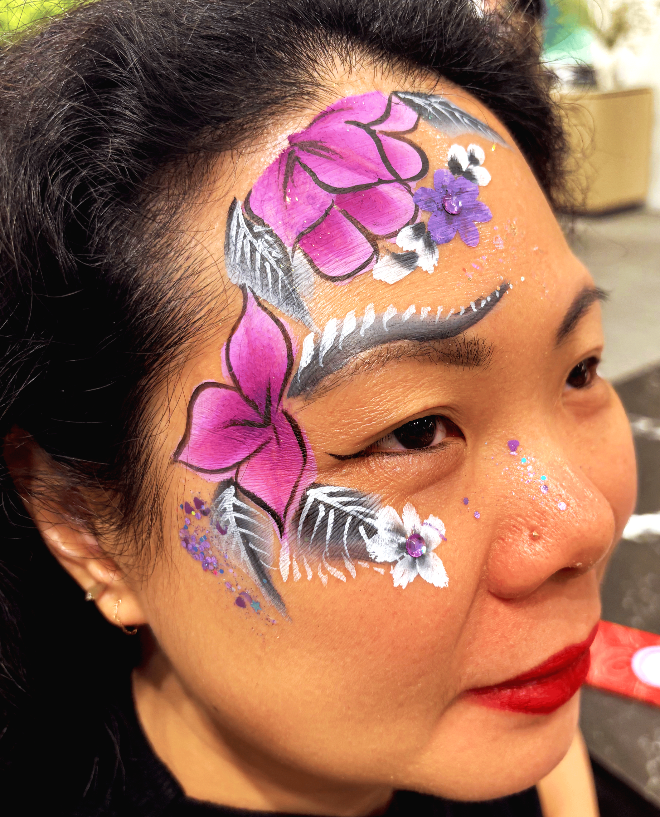 Close-up of a woman's face with colorful face paint, featuring pink, purple, and black designs, along with white accents and glitter, emphasizing her cheek, eye, and forehead.