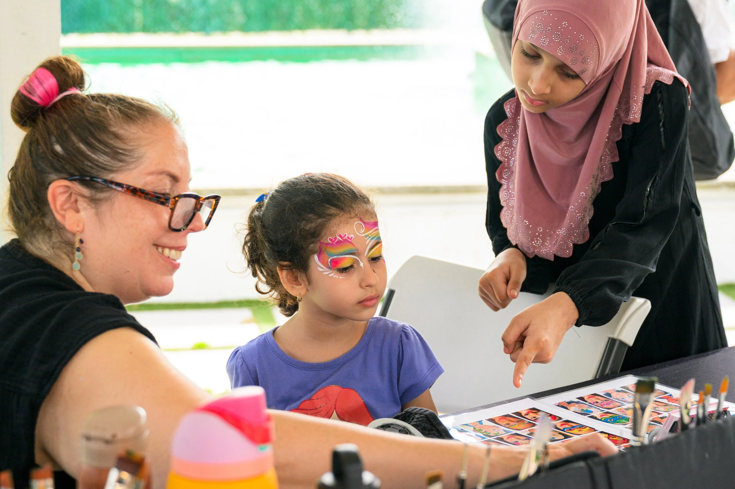 A girl being face painted by a woman with glasses sitting while a woman in a pink hijab watches