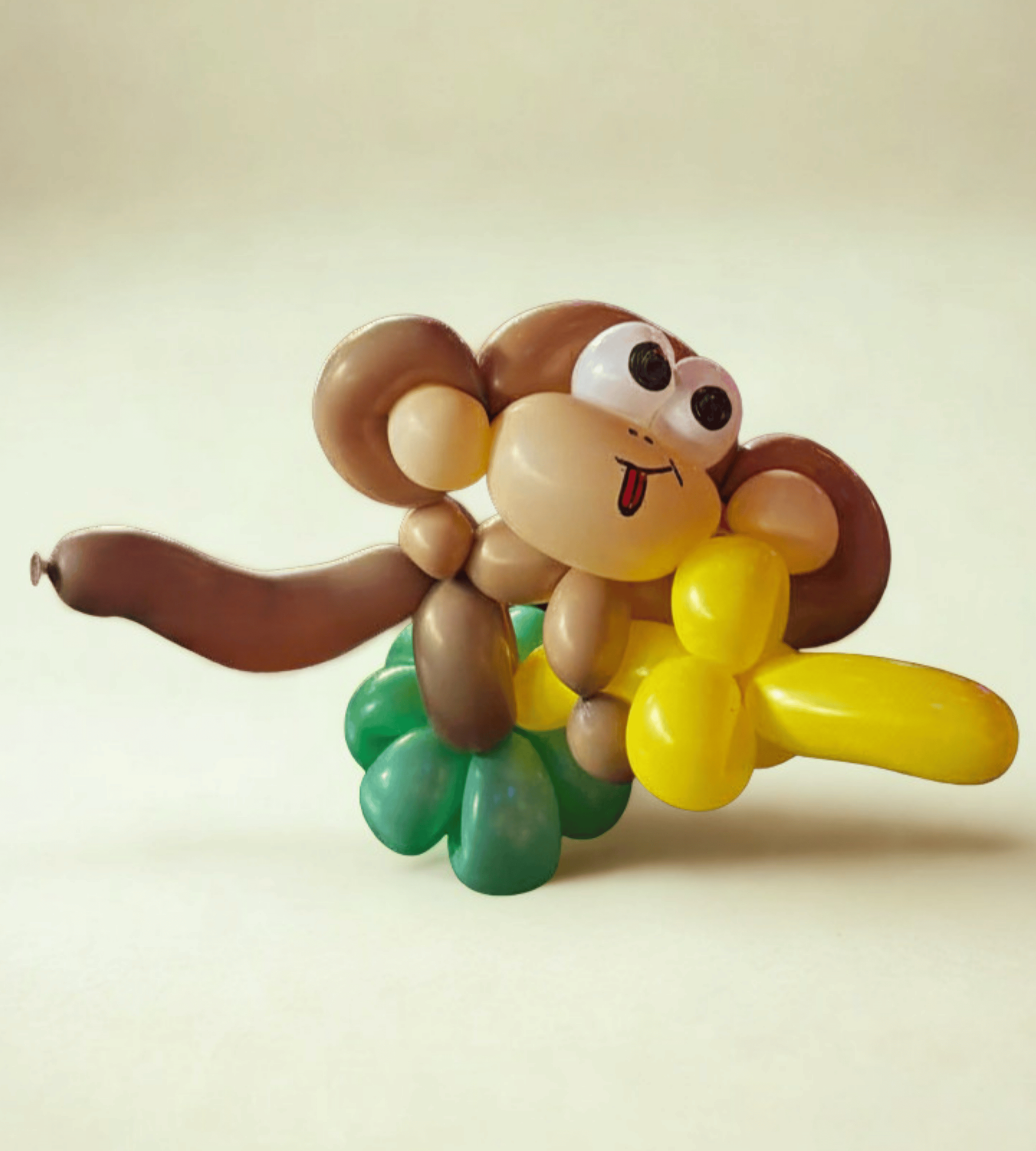 A balloon sculpture of a cute monkey holding a banana with exaggerated eyes and a tongue sticking out, set against a plain background.