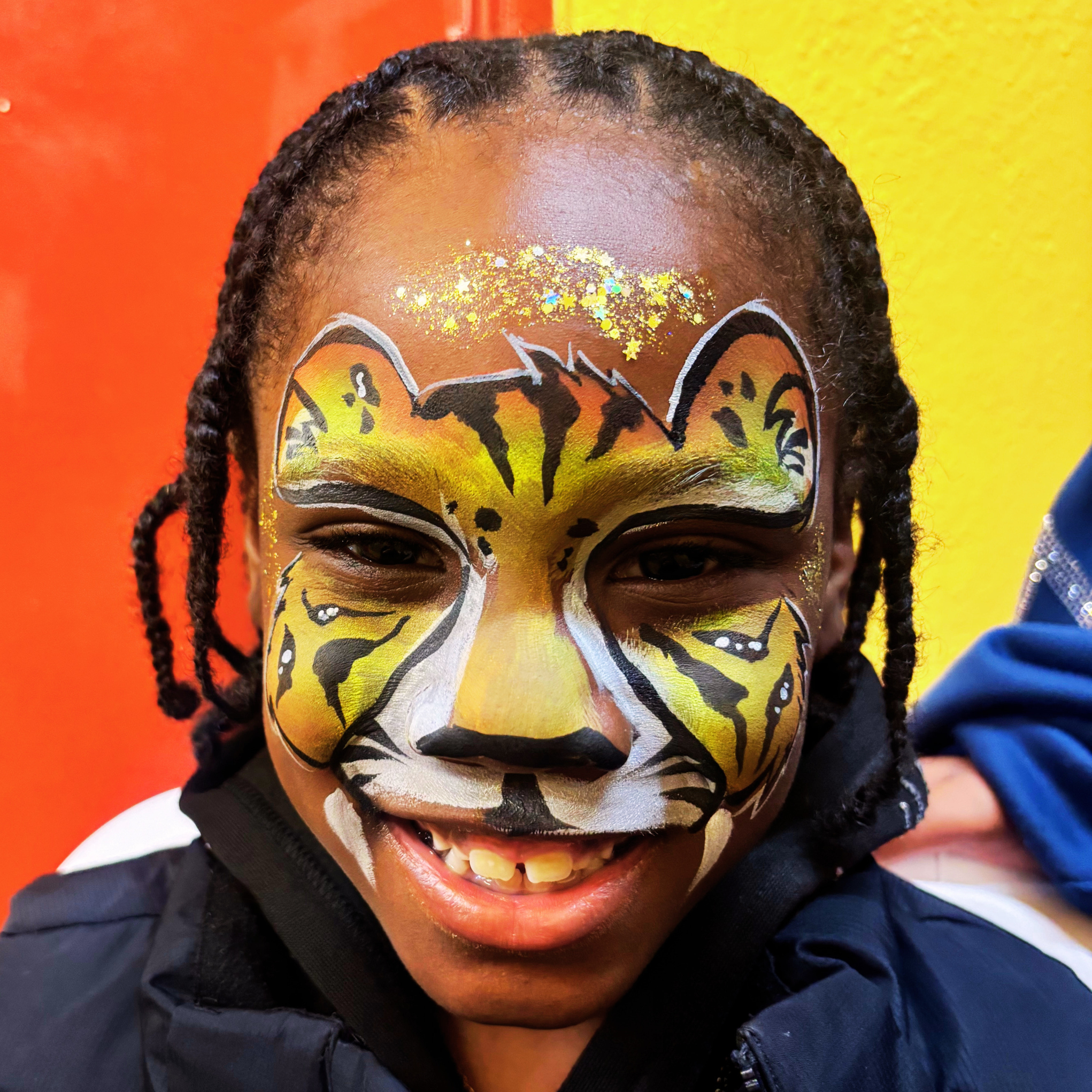 Child with face painted as a tiger smiling with eyes closed, wearing a dark hoodie, against a yellow and red background.