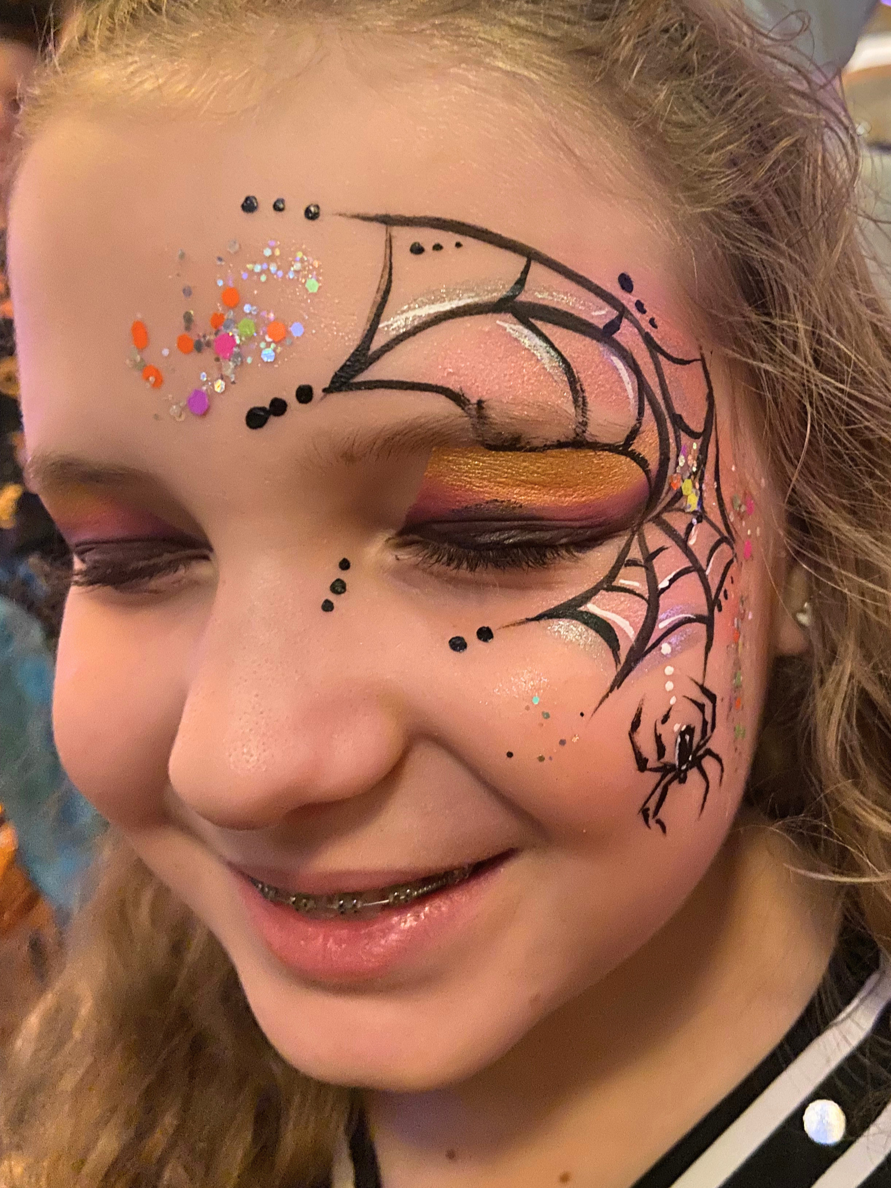 A close-up of a person's face featuring elaborate makeup with glitter and colorful accents, including a painted spider web design around the eye, gold and pink eyeshadow, and multicolored rhinestones and glitter.