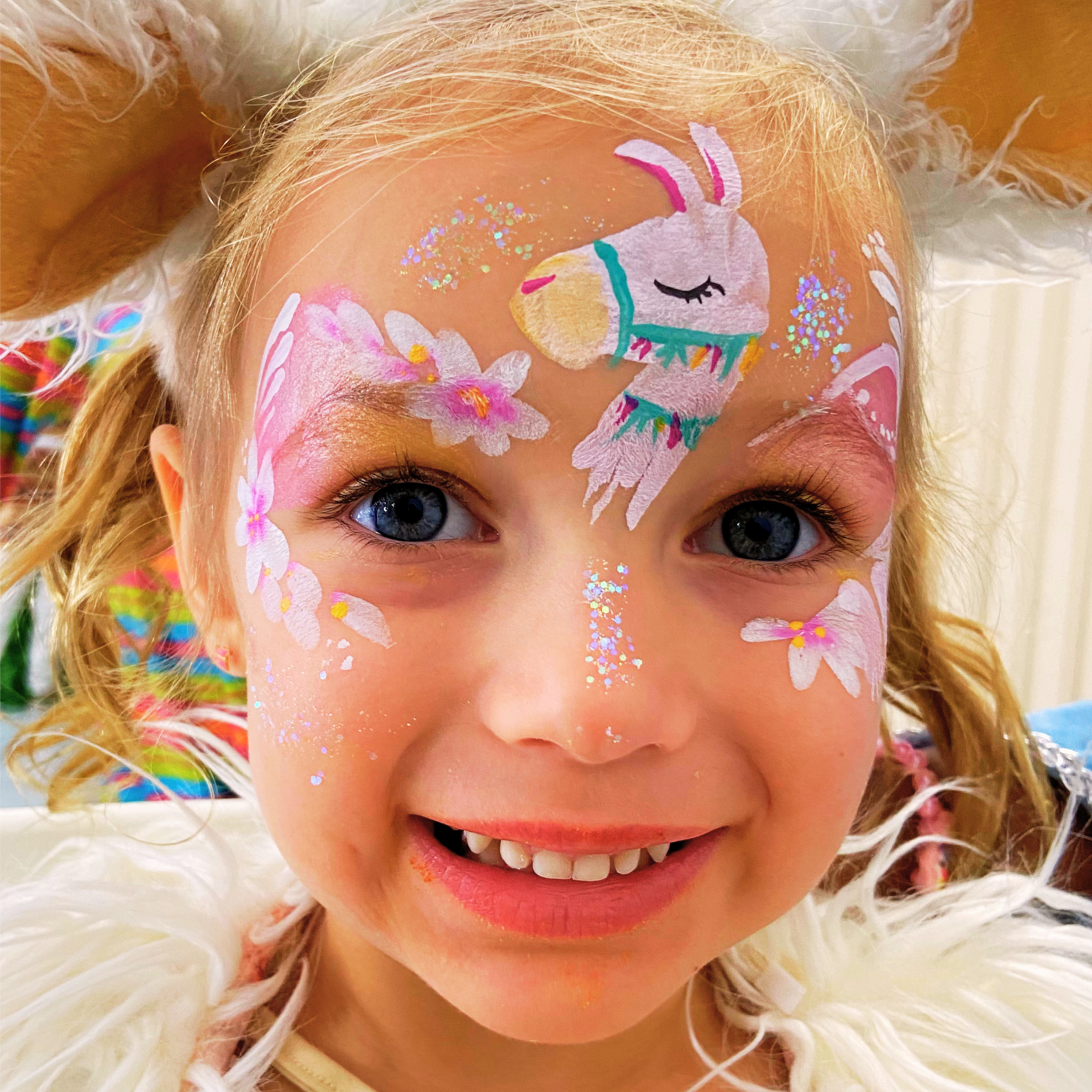 Close-up of a young girl's face with colorful llama and flower face paint, glitter, and festive makeup, with her eyes wide open and a playful smile.