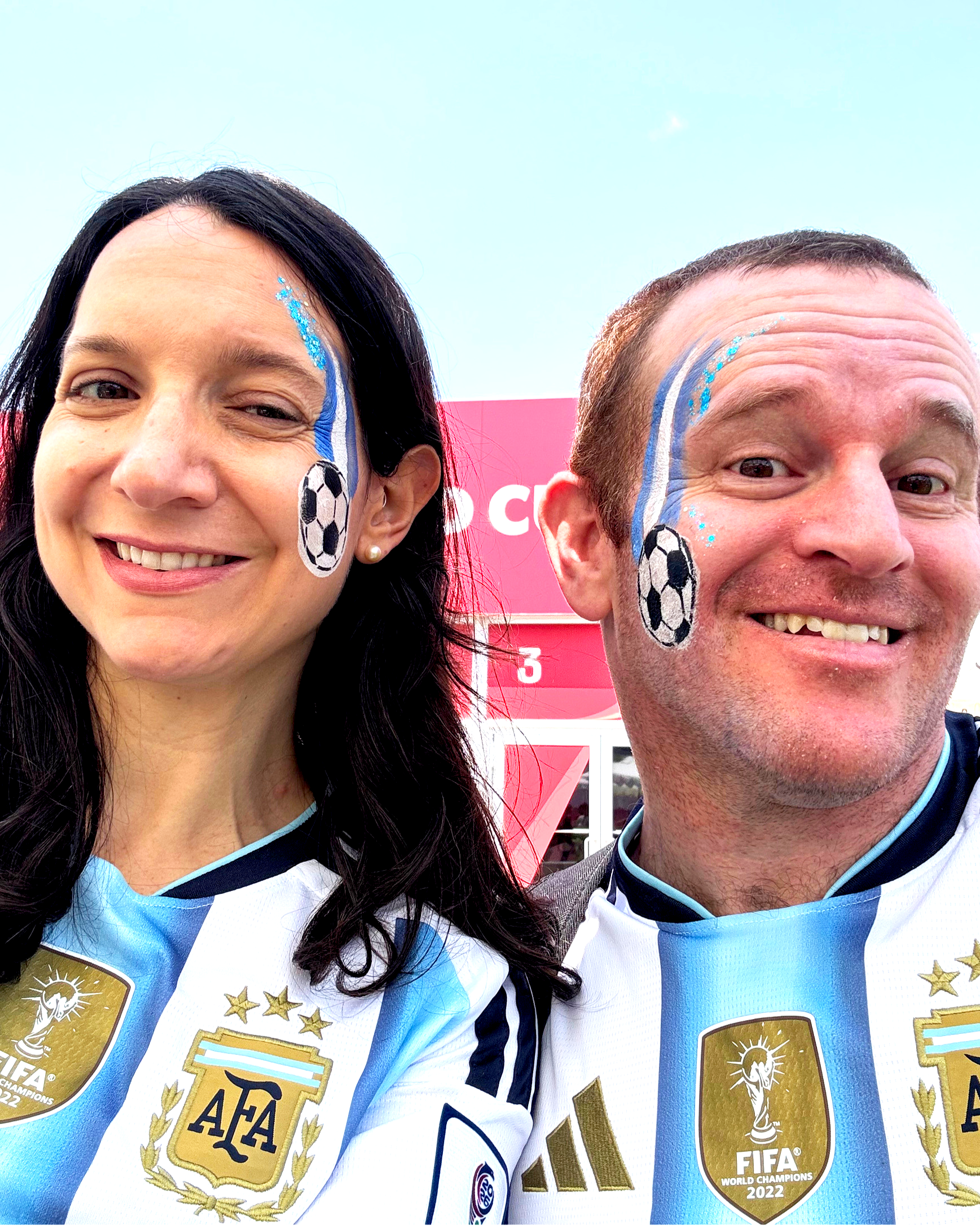 Close-up of two smiling soccer fans with face paint and team jerseys, celebrating at the 2022 FIFA World Cup, with a pink background and part of a sign visible.