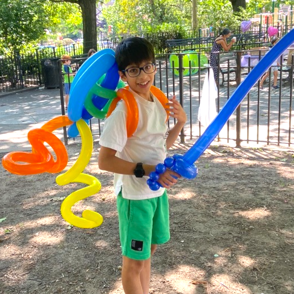 A boy smiling and holding a balloon sword at a park, with a colorful balloon jetpack, and other children playing in the background.