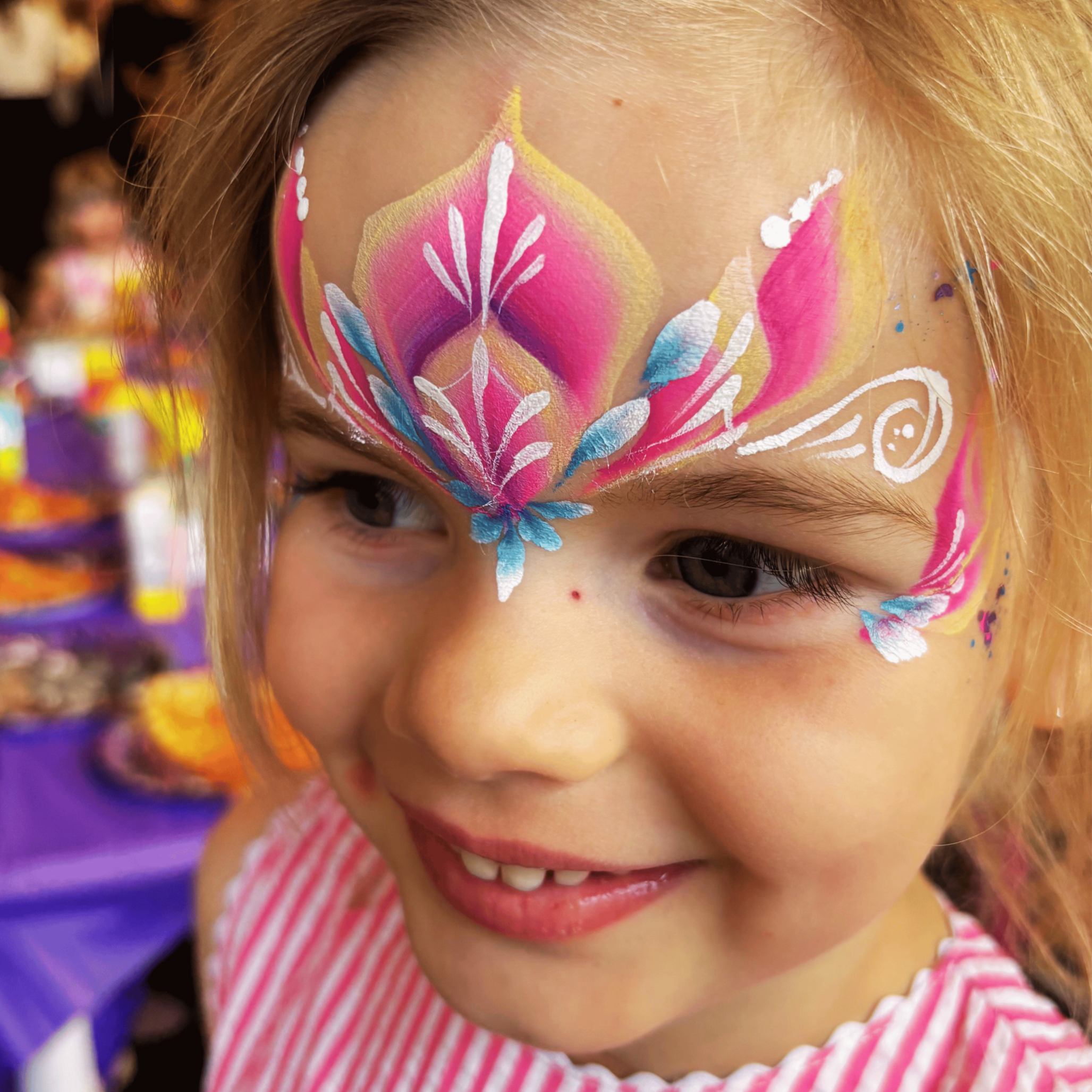Close-up of a young girl with face paint in bright colors, mostly pink, white, blue, and yellow, and wearing a pink and white striped shirt. She is smiling and tilting her head slightly downward.