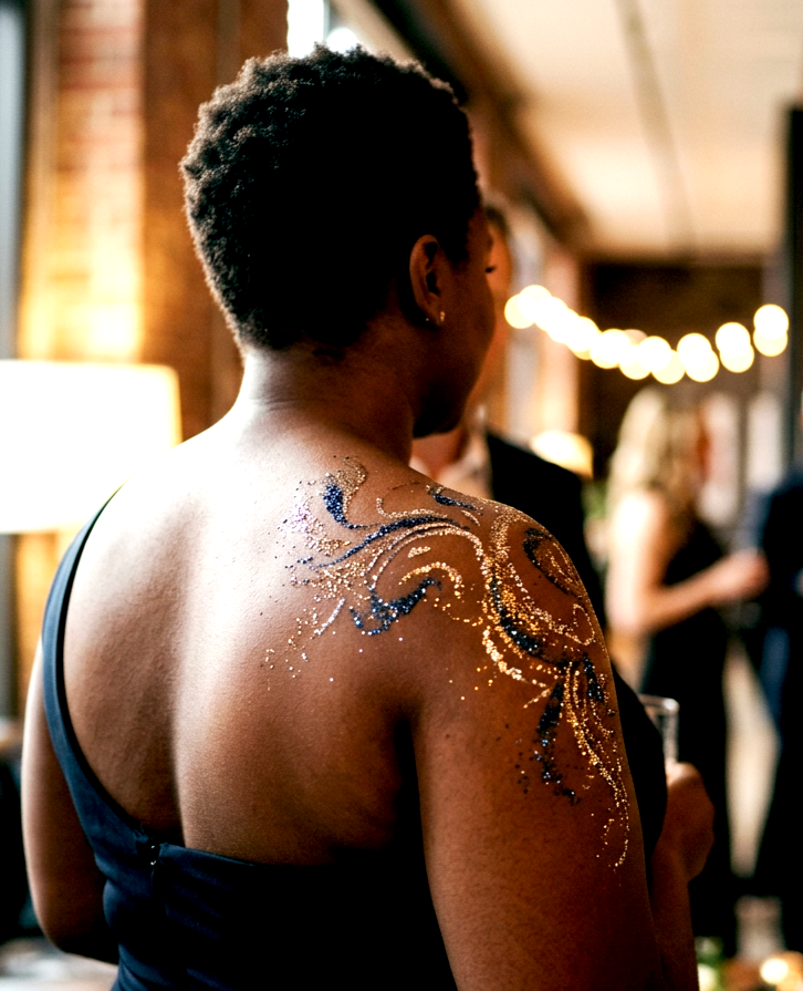 A woman with short curly hair and dark skin, wearing a sleeveless black top, has a colorful glitter tattoo on her right shoulder at a social gathering or party.