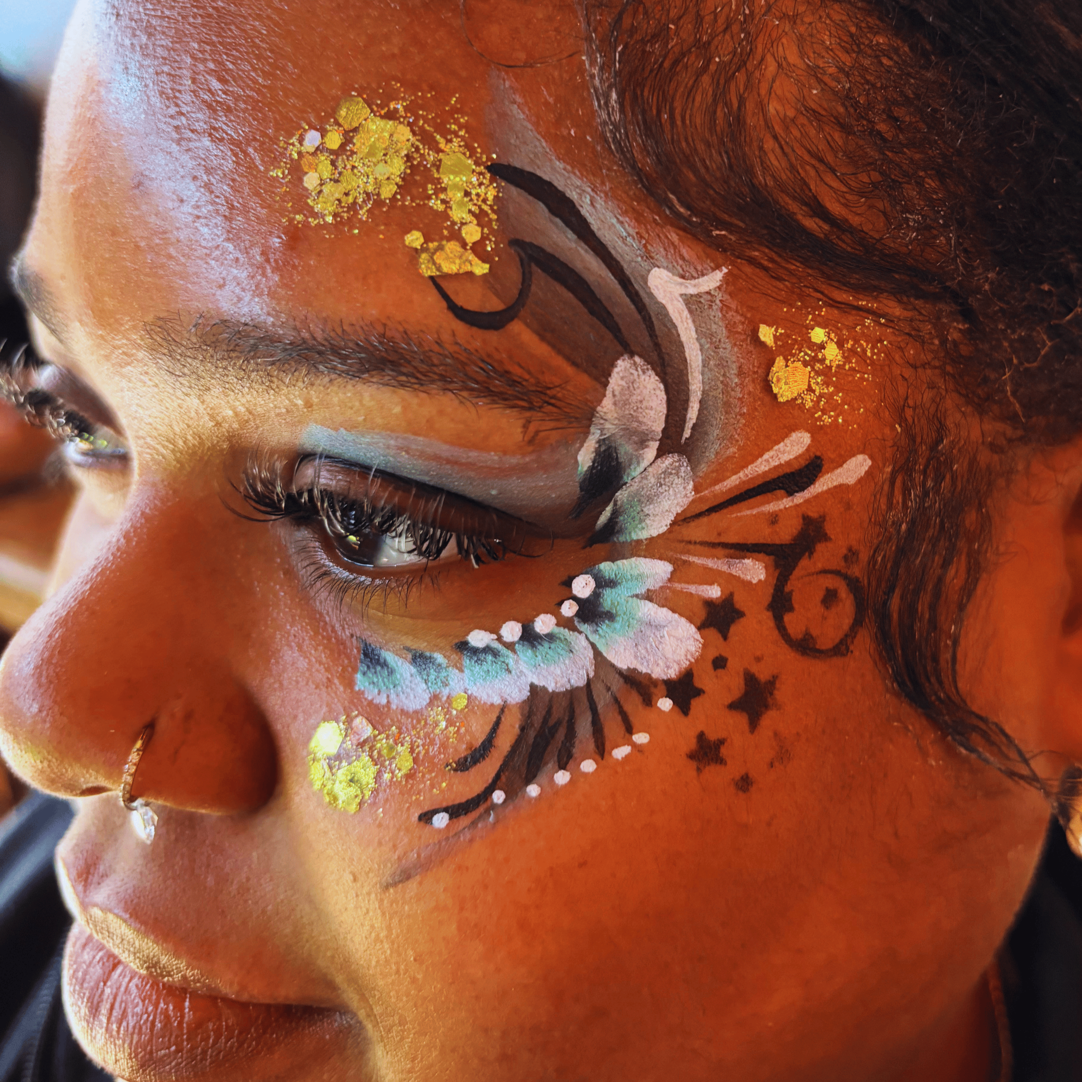 Close-up of a person's face with colorful, artistic makeup and face paint, featuring yellow, black, white, and light blue colors, along with black lines and star shapes around the eyes and cheeks.
