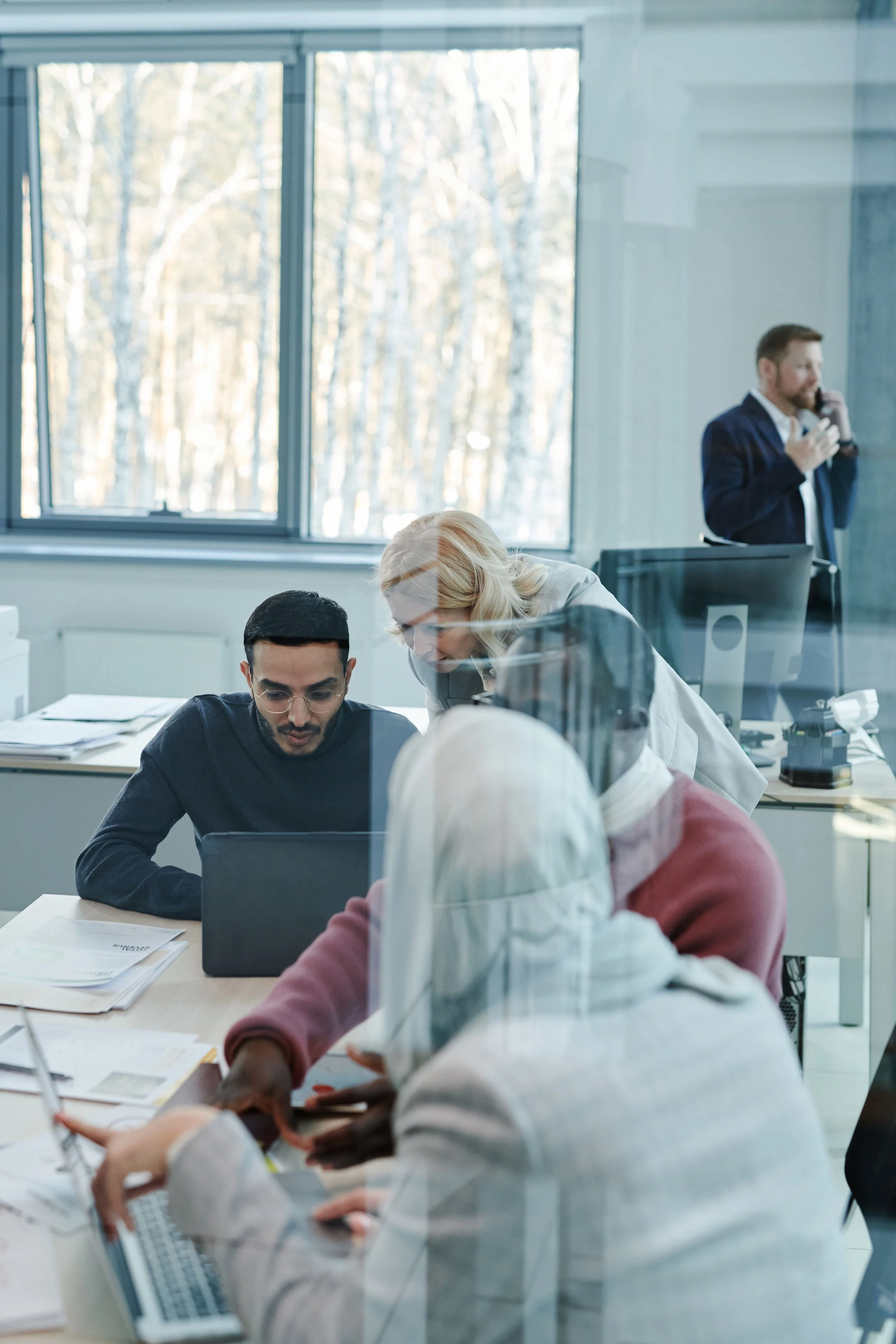 People working together in an office, with a man at a computer in the foreground and a woman leaning over explaining something. A man in a suit speaking on the phone in the background.